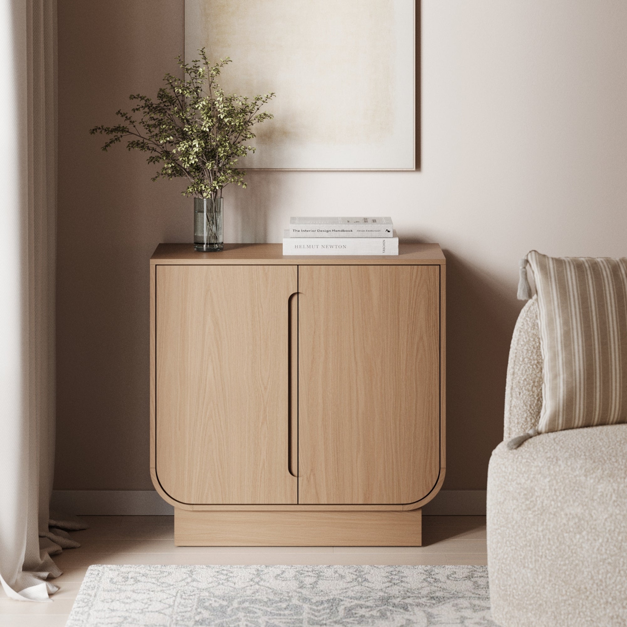 A Nathan James Wood Veneer Arched Accent Cabinet in Raw Oak with rounded edges stands against a beige wall, topped with a glass vase of green branches and three books. A striped cushion and light rug complete the scene.
