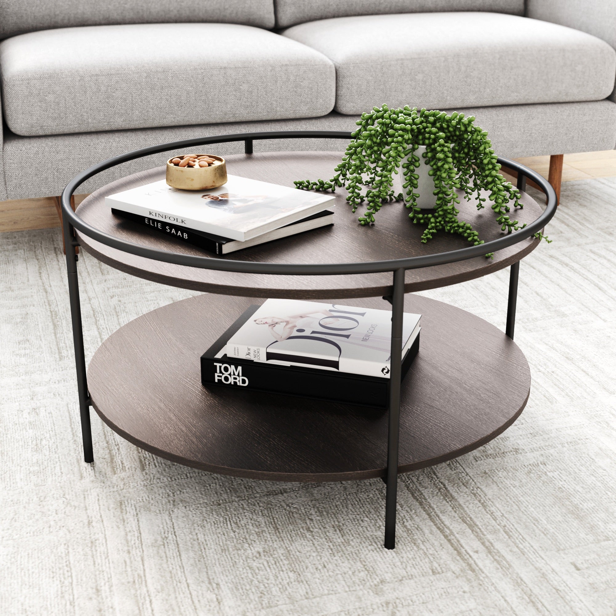 A round, dark wood coffee table with black metal legs holds books, a small potted plant, and a bowl. A gray sofa is in the background, and the table sits on a light textured rug.
