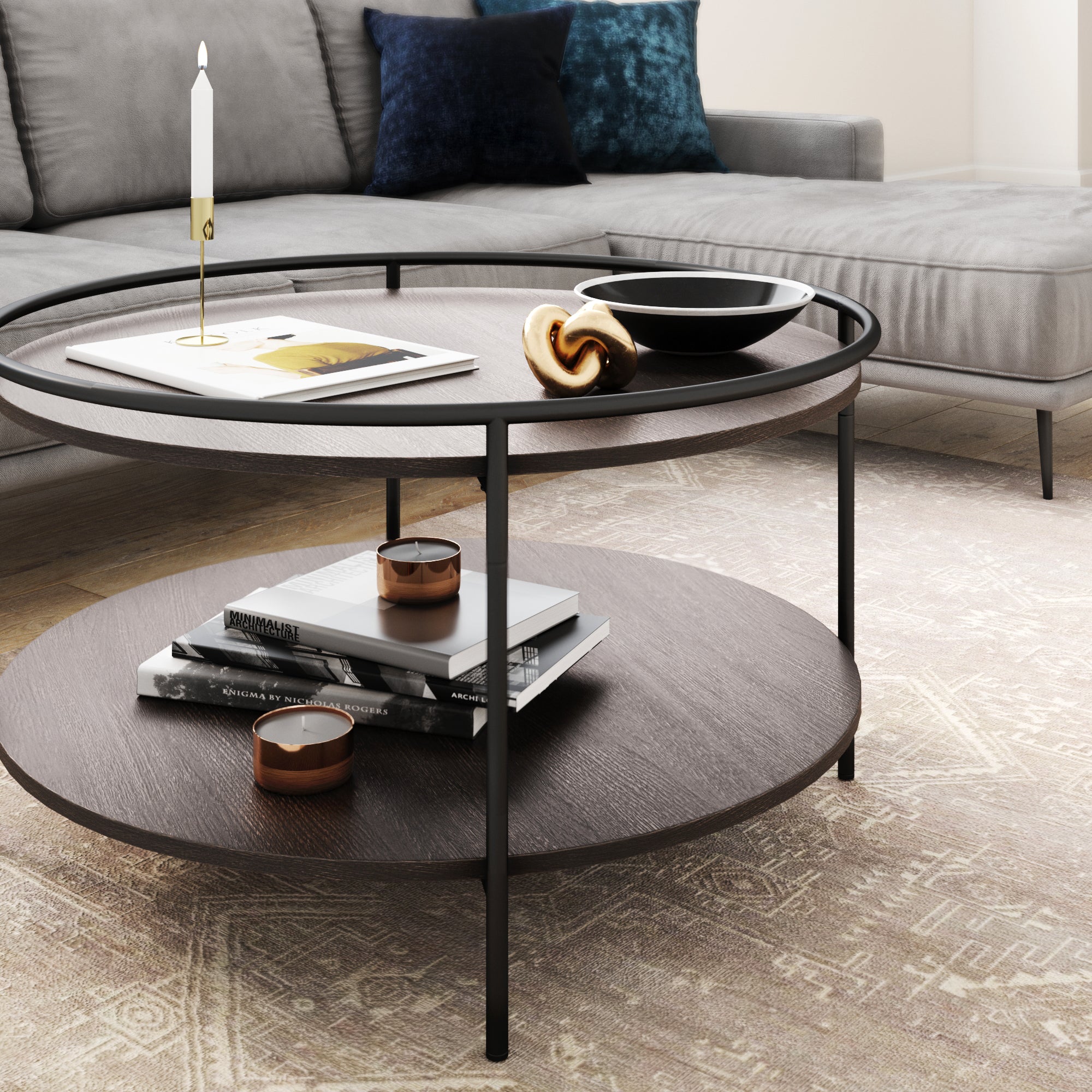 A round wooden coffee table with a black metal frame holds a candle, book, decorative bowl, and pastry. The lower shelf has books and small brown containers. A gray sectional sofa and patterned rug are in the background.