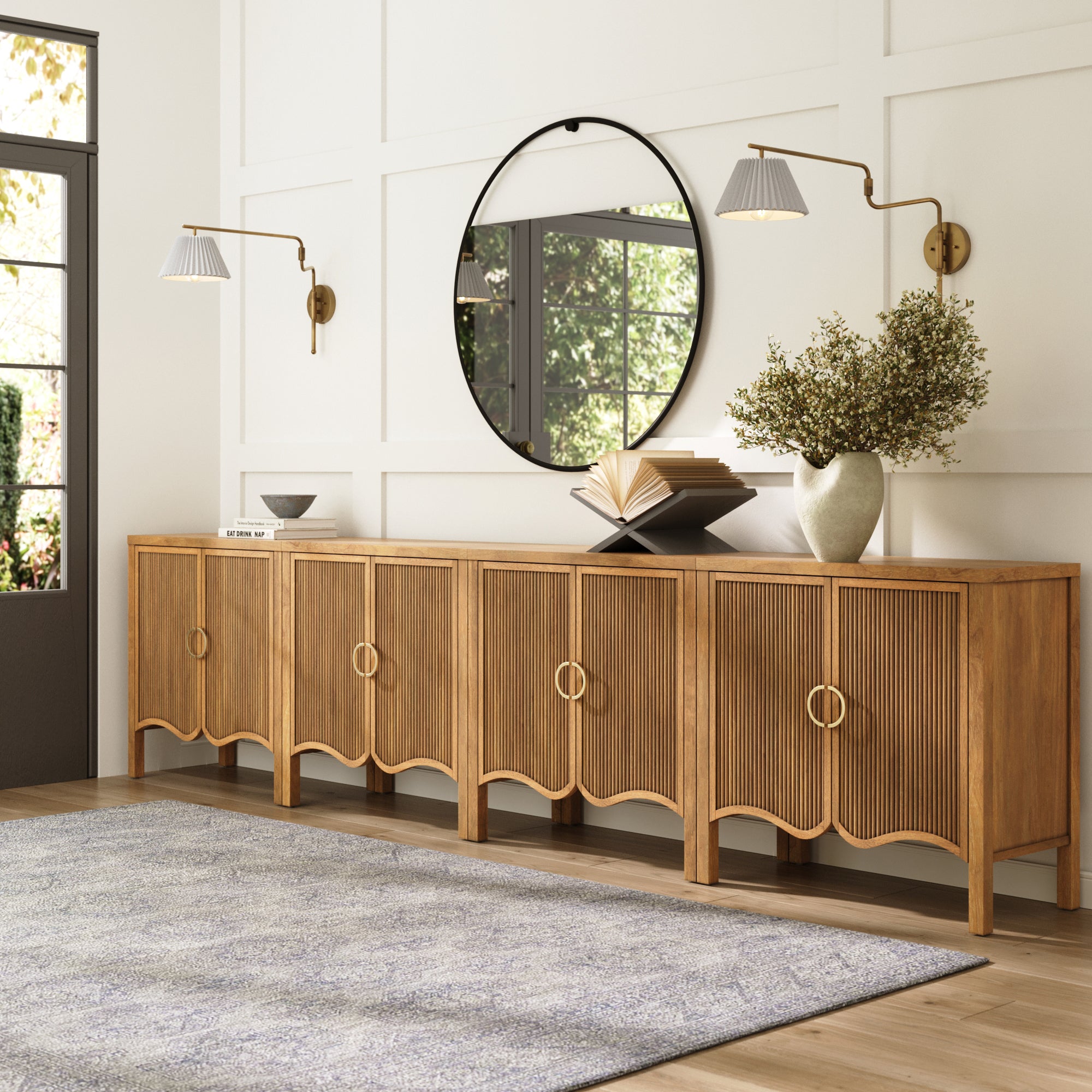 A modern living room showcases the Nathan James Transitional Fluted Large Credenza Cabinet (Set of 4), round mirror, sleek wall sconces, flower vase, open books, and a gray rug on light wood floors with sunlight streaming in.