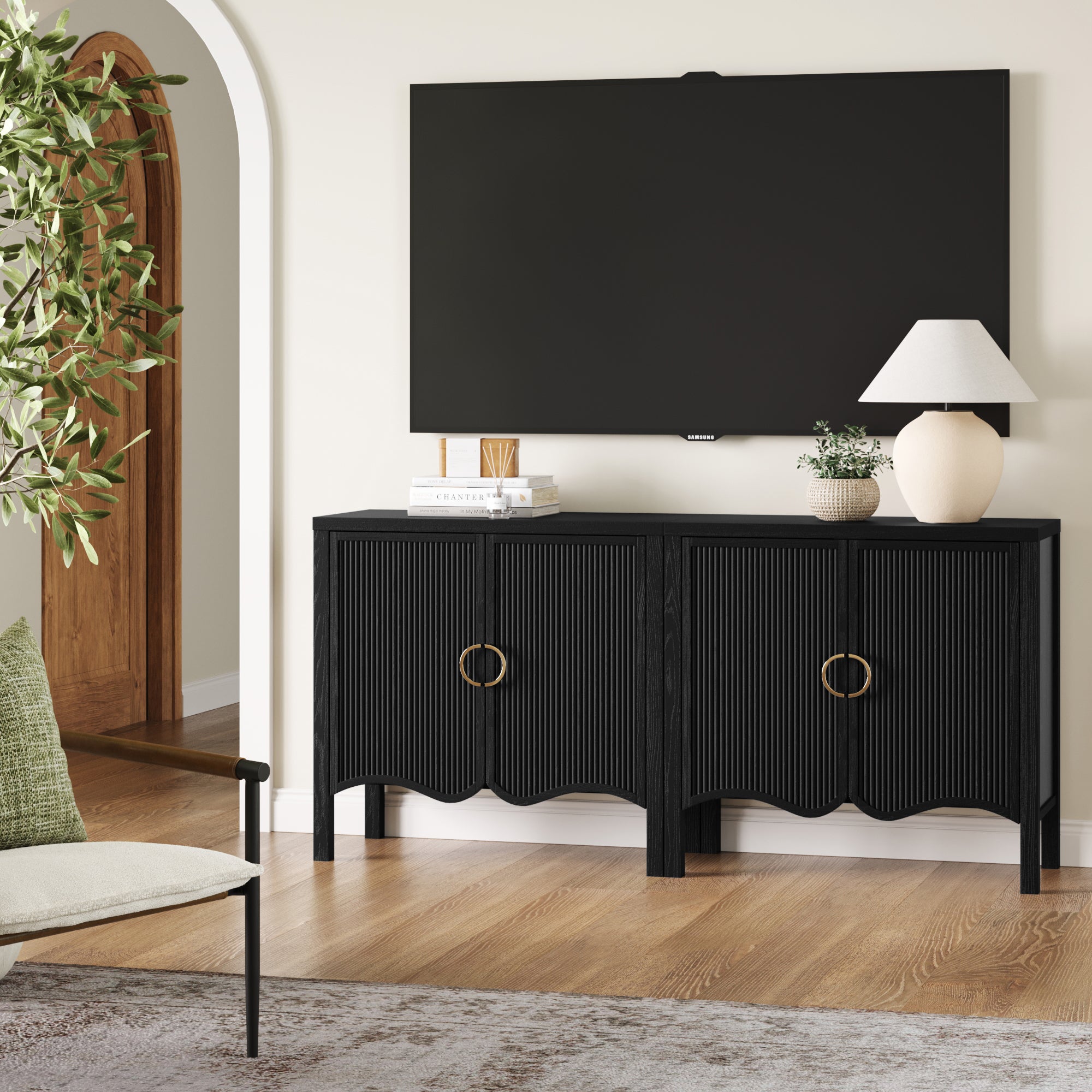 A modern living room features a wall-mounted TV above the Nathan James Transitional Fluted Cabinet or TV Stand in Black Oak (Set of 2), with gold ring handles and fluted doors. Decor includes a white lamp, books, a plant, chair, and potted tree nearby.