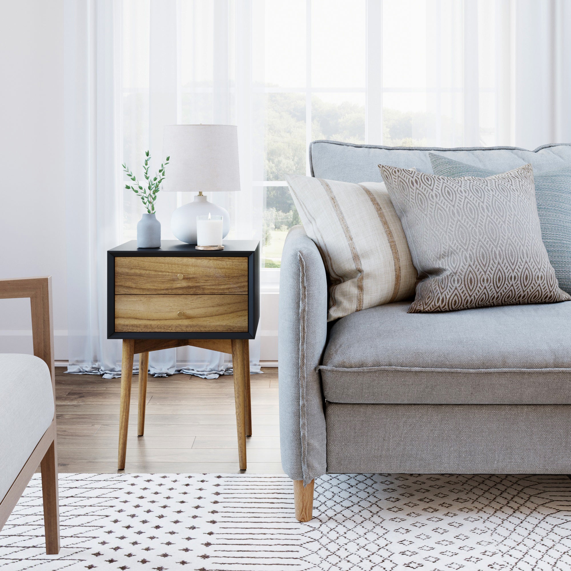 A modern living room with a light gray sofa, patterned cushions, a geometric rug, and a wooden side table holding a lamp, a small plant, and a book, set against large windows with sheer white curtains.