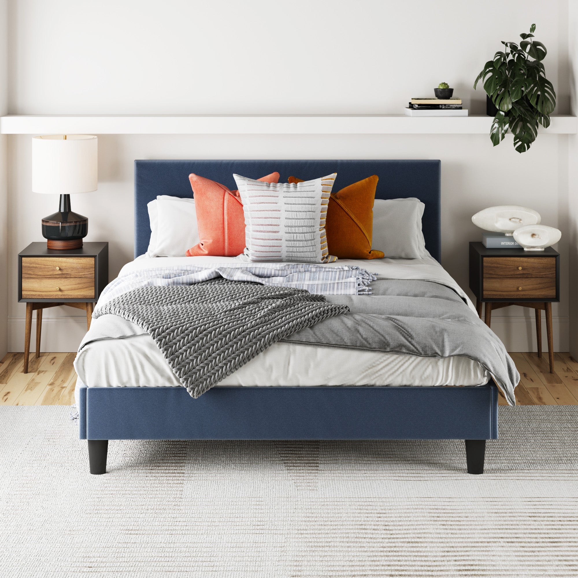 A modern bedroom featuring a blue upholstered bed, colorful pillows, gray blanket, and Nathan James Wood Mid-Century Nightstands in black with lamps and books, plus a potted plant on the right atop a light rug and wooden floor.