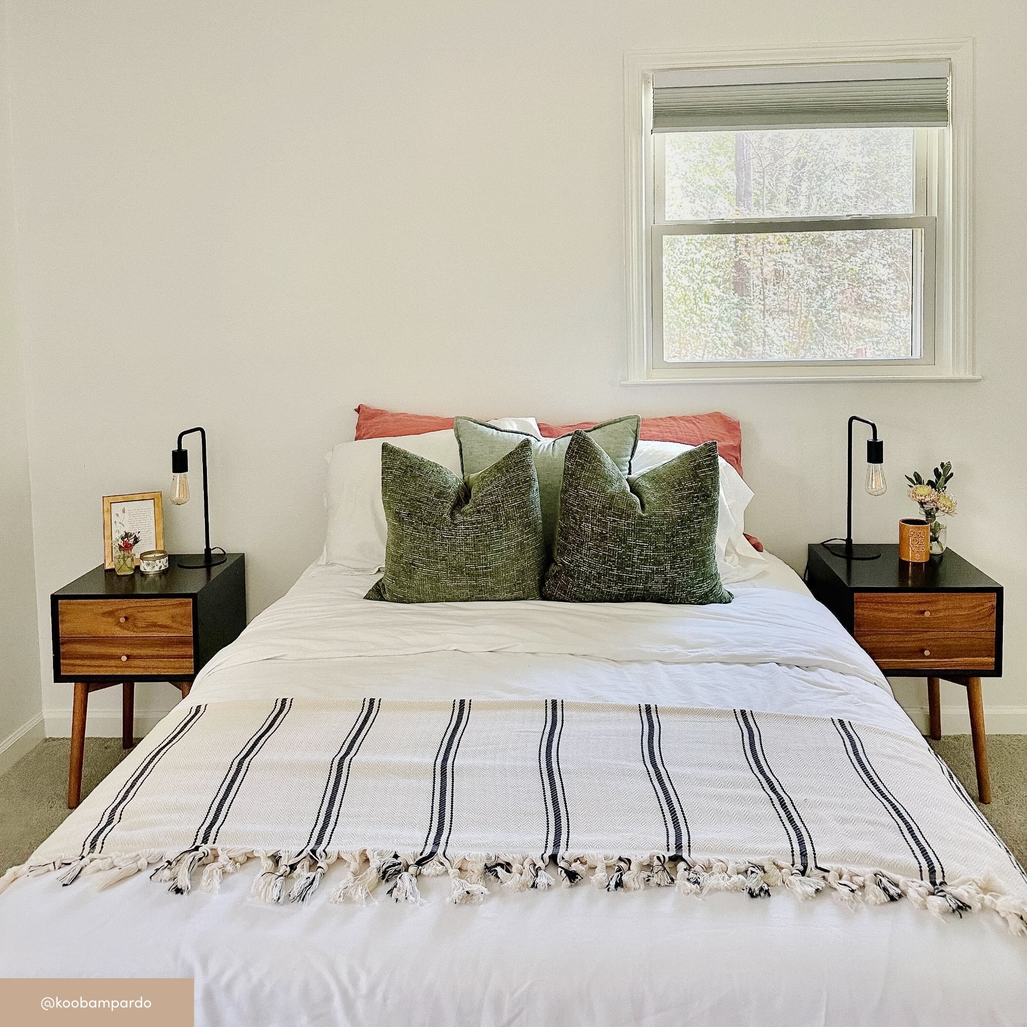 A neatly made bed with a white comforter, green throw pillows, and a striped blanket. Two black and wood nightstands with lamps, a framed picture, and decor sit on each side. A window is above the bed on a white wall.