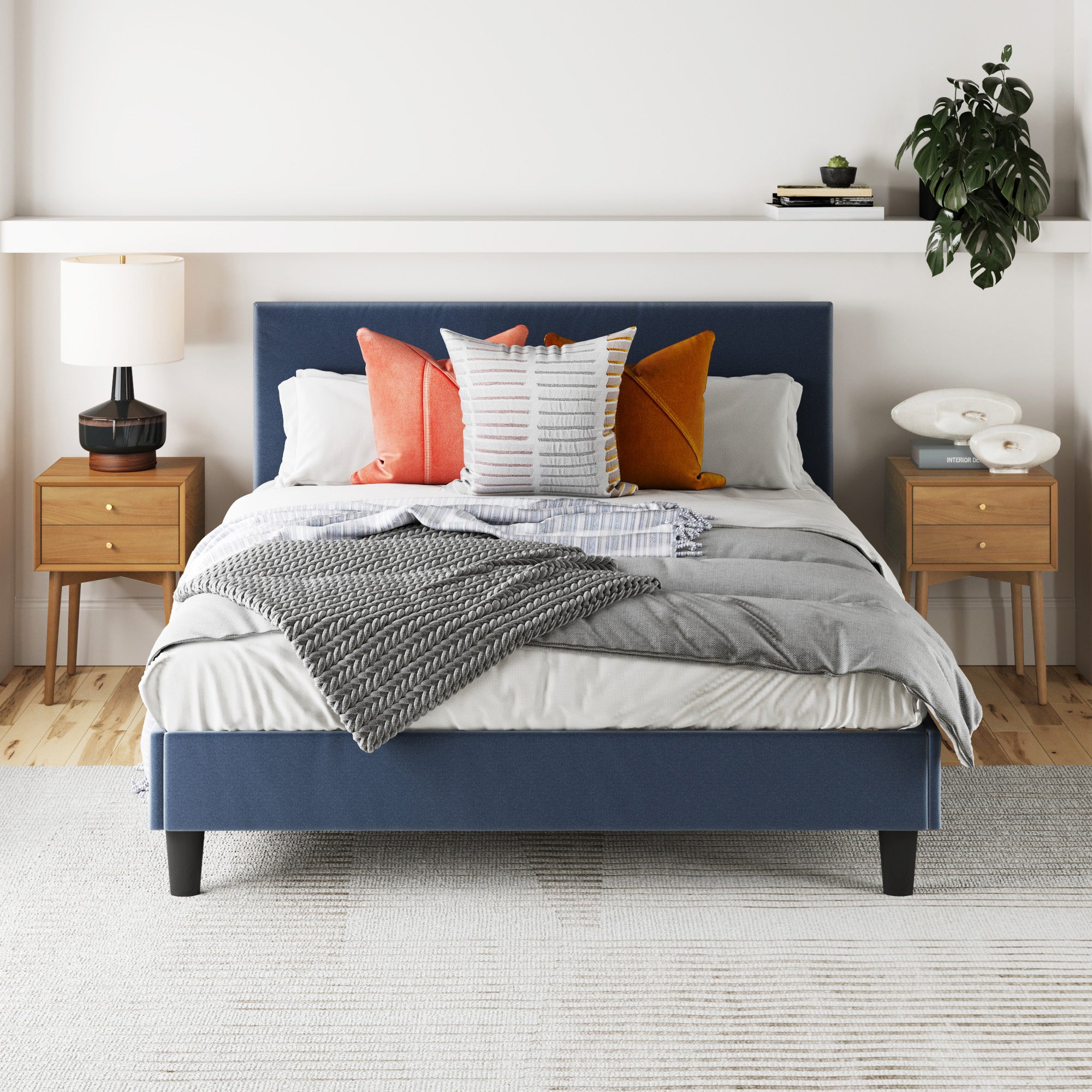 A modern bedroom featuring a blue upholstered bed with gray and white bedding, colorful pillows, and Nathan James Wood Mid-Century Nightstands Acacia (Set of 2), plus vintage brass lamps and a plant on a shelf above the bed.