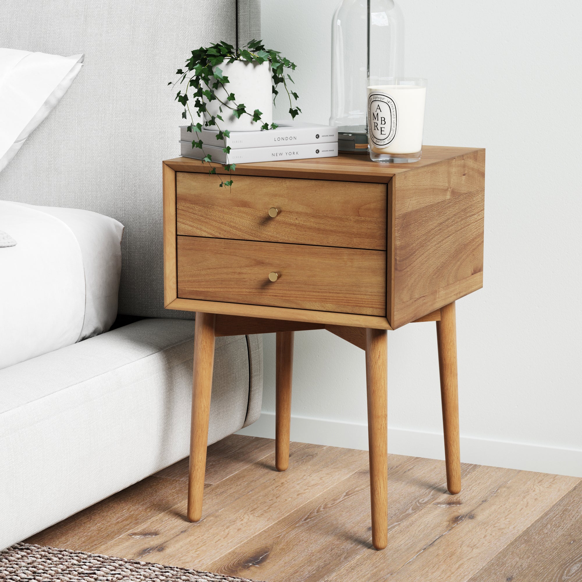 A Nathan James Wood Mid-Century Nightstand Acacia with vintage brass hardware and two drawers stands by a bed, topped with a trailing plant, books, a candle, and a glass container. The room has wood floors and white walls.