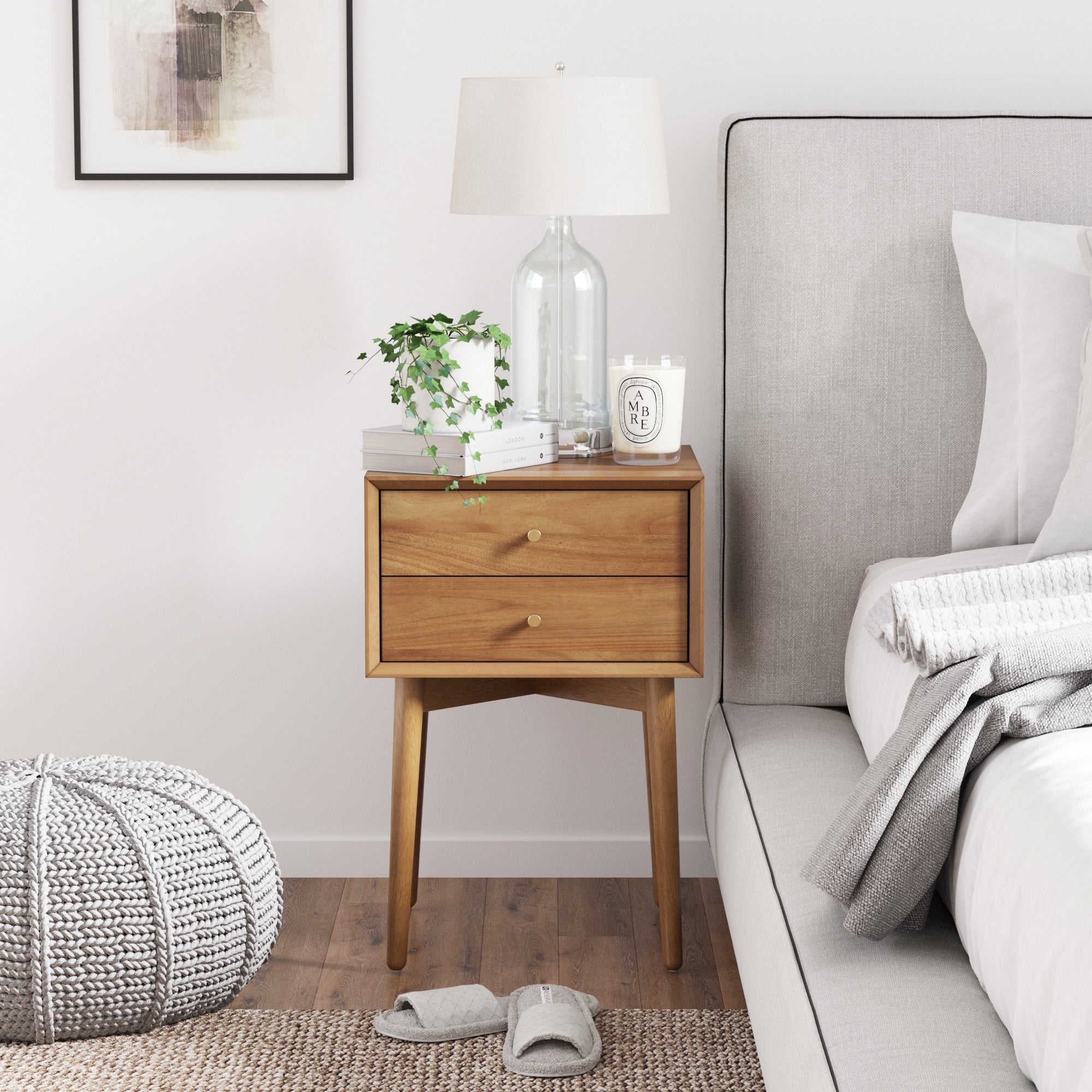The Nathan James Wood Mid-Century Nightstand Acacia with vintage brass hardware and two drawers holds a glass lamp, potted plant, candle, and book beside a bed with a gray upholstered headboard. Slippers and a knitted pouf sit on the wood floor.