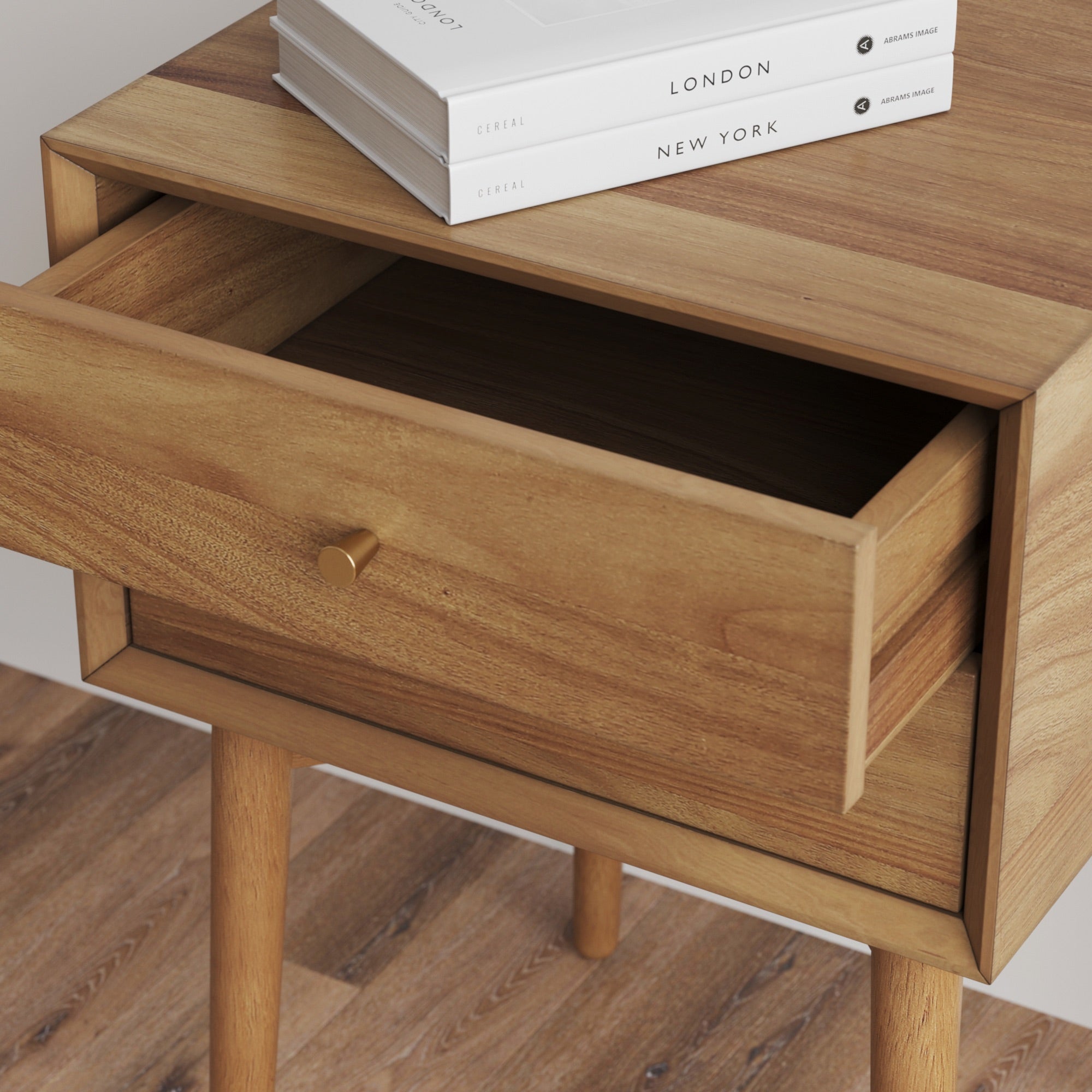 A wooden nightstand with an open drawer, gold knob, and two stacked books titled London and New York on top, placed on a wooden floor.
