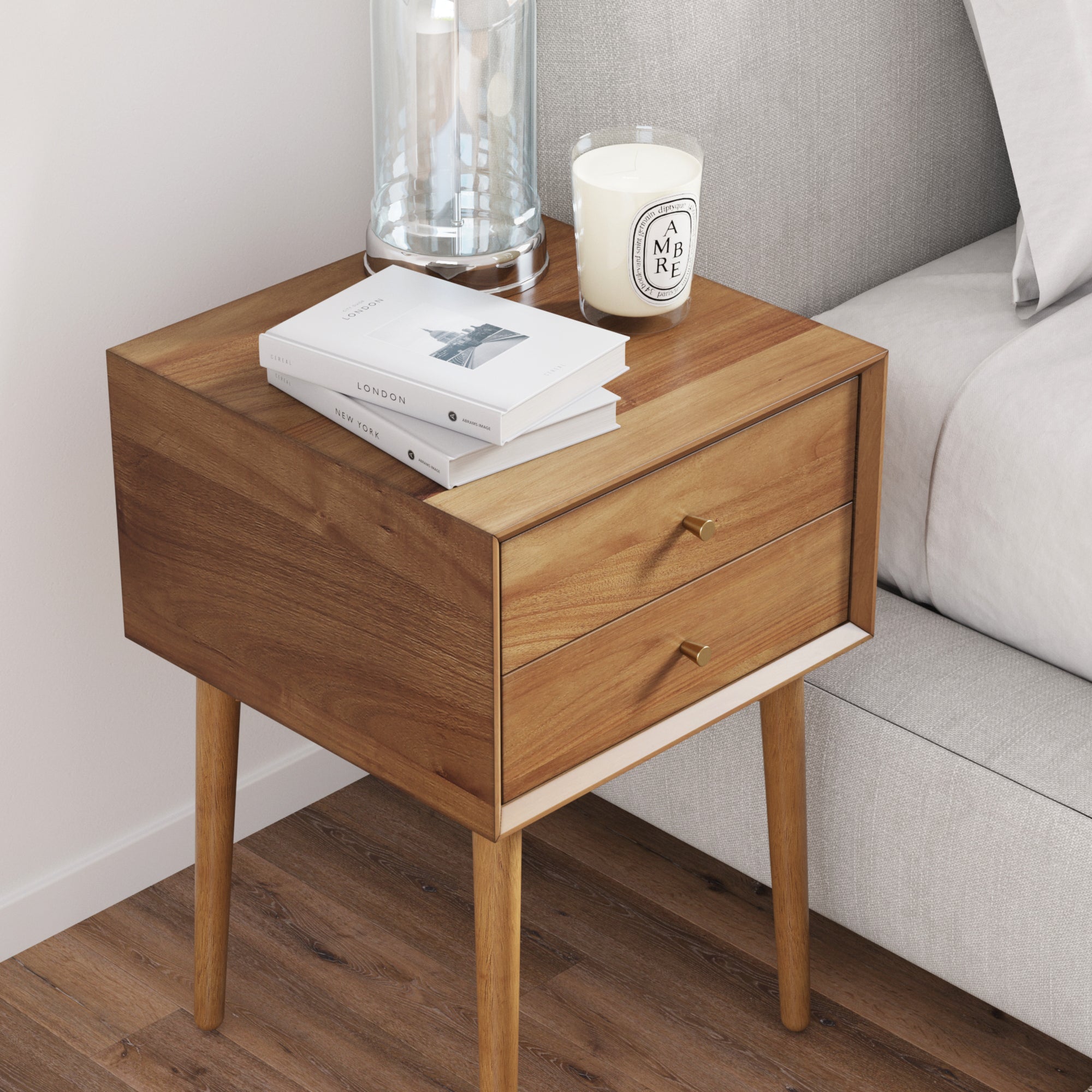A wooden nightstand with two drawers stands by a gray upholstered bed. On top are a glass lamp, a white candle, and two stacked books. The nightstand has gold drawer pulls and tapered legs on a wooden floor.