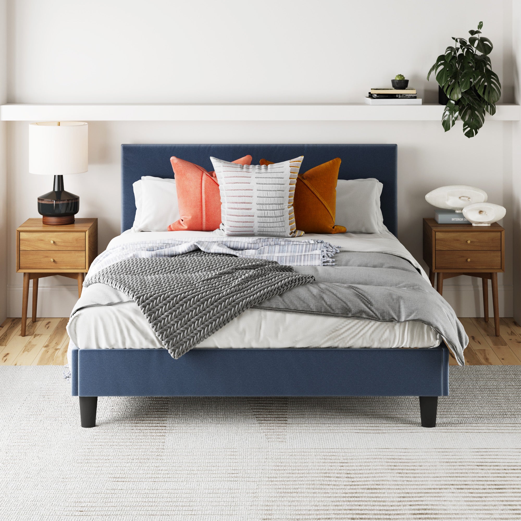 A neatly made bed with a blue upholstered frame, layered gray and white bedding, and colorful accent pillows sits between two wooden nightstands with lamps in a bright, modern bedroom. A plant hangs on the right wall shelf.