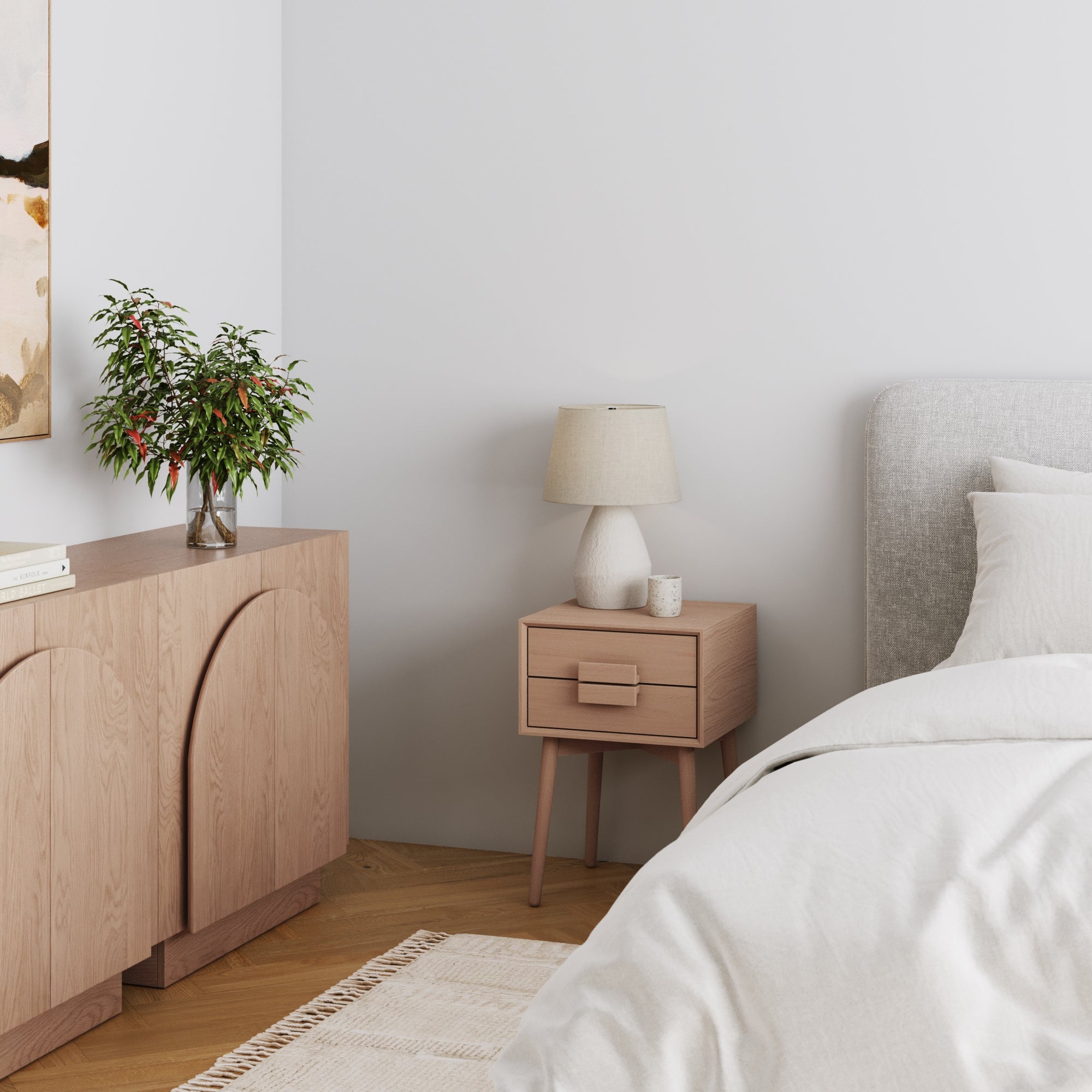A bedroom featuring Nathan James’s Mid-Century Wood Handle Nightstand in Raw Oak, a matching dresser, beige lamp, plant in vase, gray upholstered bed with white bedding, cream rug on wood floors, and white walls.