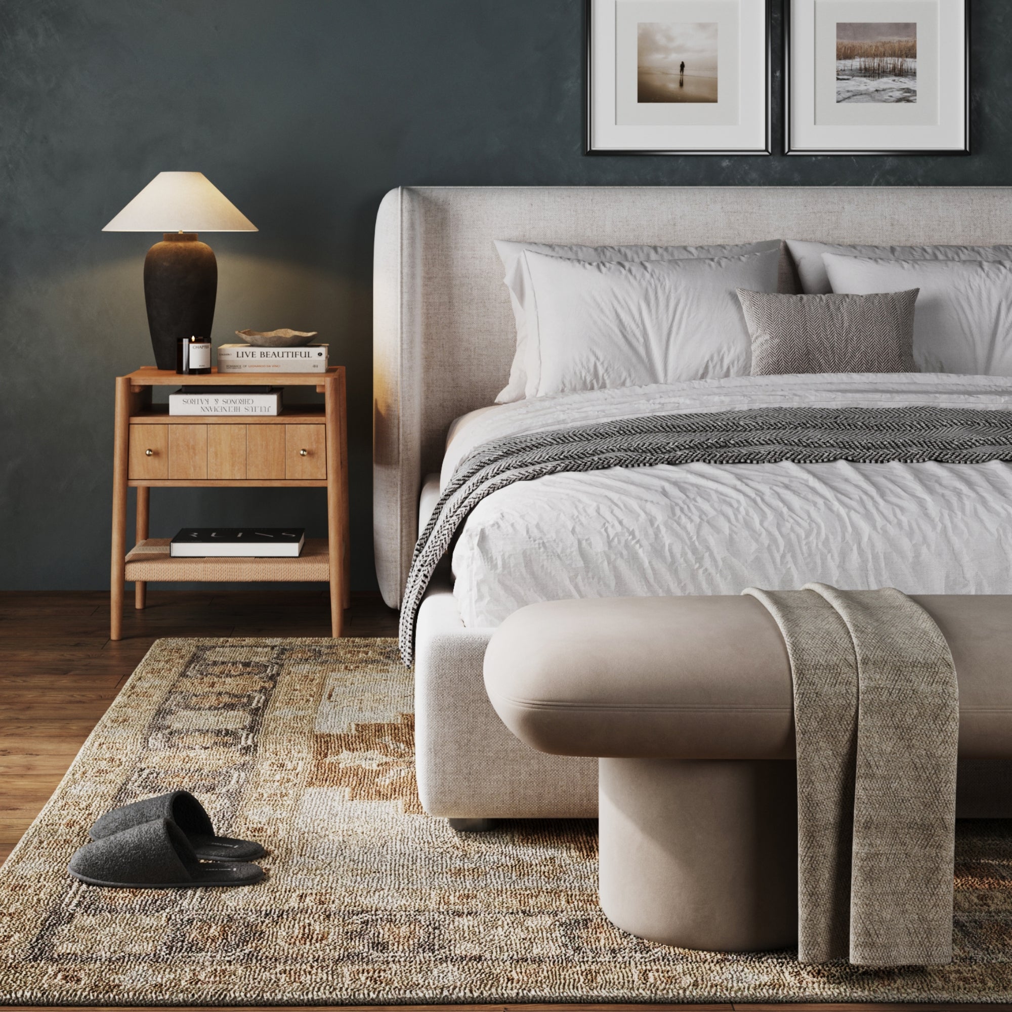 A modern bedroom features a plush beige bed, a Nathan James Wood Nightstand with Drawer & Seagrass Storage in light brown holding a lamp and books, soft rug, gray slippers on the floor, and neutral decor with framed photos on a dark wall.