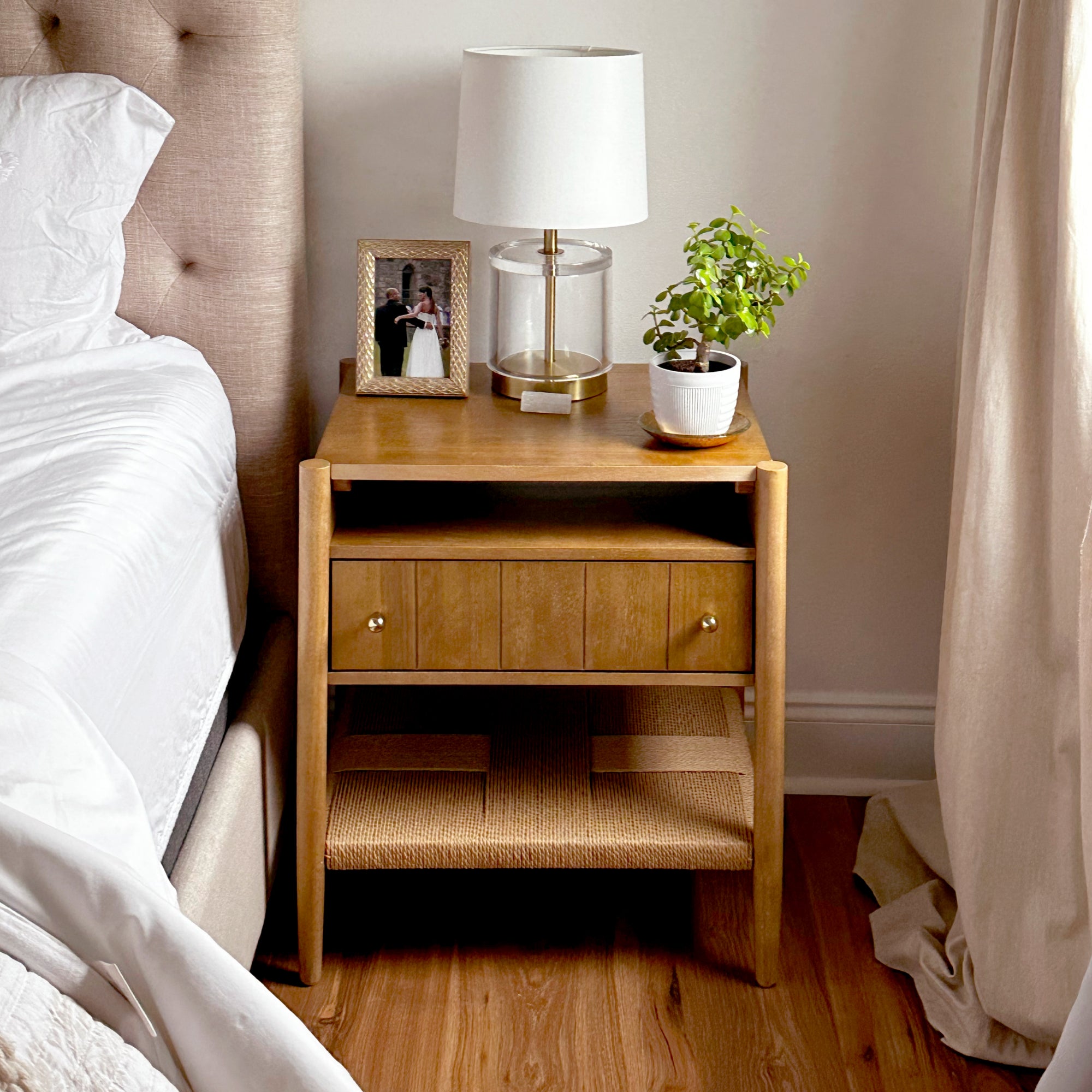 A wooden nightstand beside a bed holds a framed photo, a lamp with a white shade, and a small potted plant. The nightstand has a drawer and lower shelf. Beige curtains and bedding complete the cozy bedroom scene.