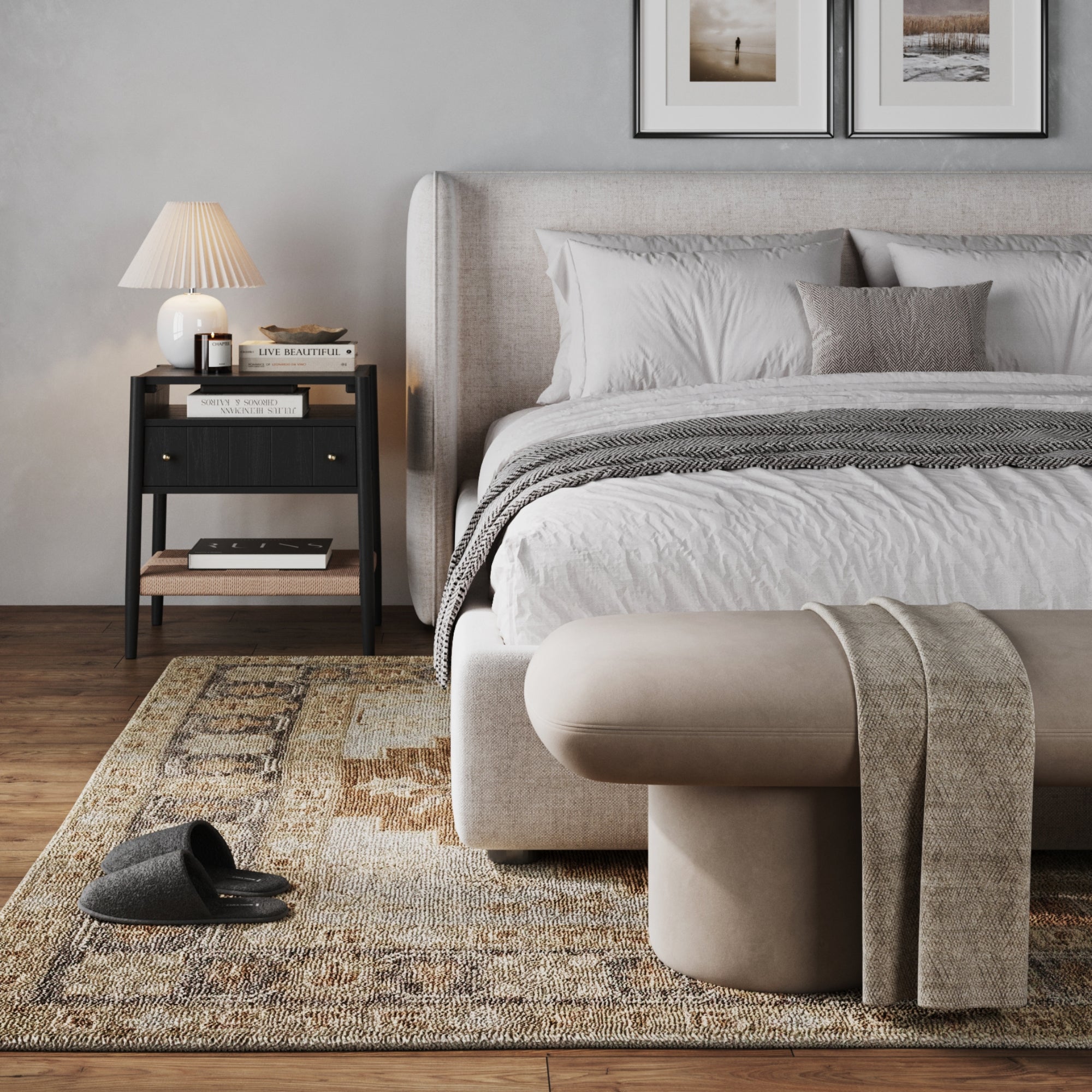 A cozy bedroom with a large upholstered bed, neutral bedding, beige bench, patterned rug, framed art, gray slippers on wood floors, and a Nathan James Wood Nightstand with Drawer & Seagrass Storage in Black Oak holding a lamp and books.