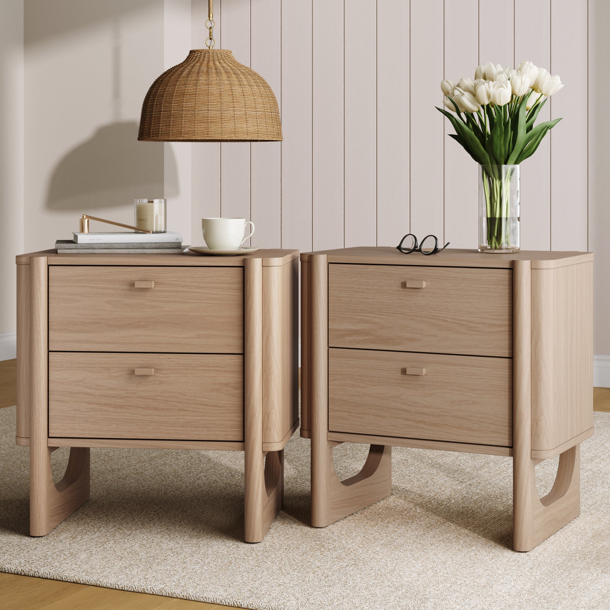 The Nathan James Mid Century Modern 2-Drawer Solid Wood Nightstands in Raw Oak (set of 2) sit on a beige rug, one topped with a vase of white tulips and glasses, the other with a tray holding a candle, cup, and book. A woven pendant light hangs above.