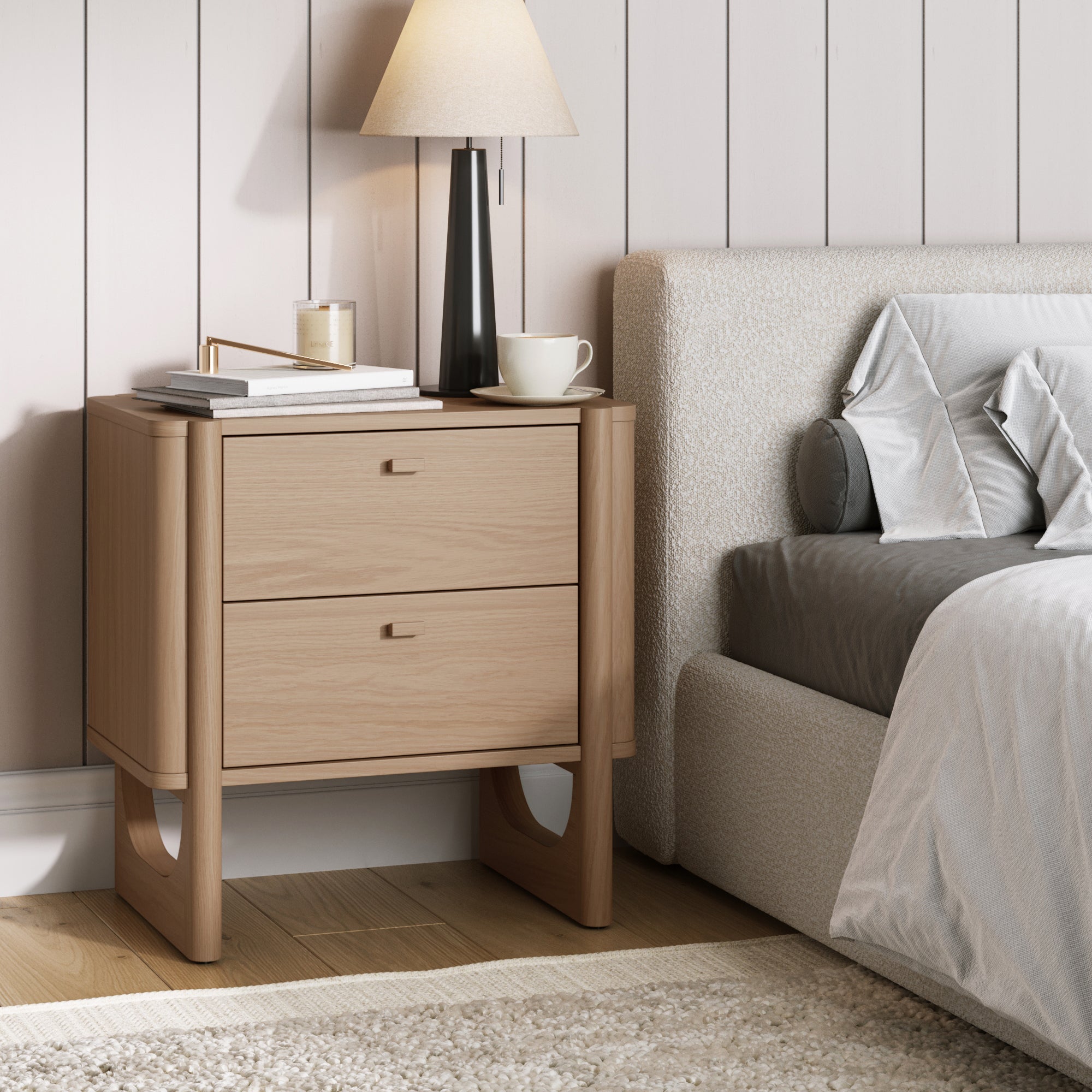The Nathan James Mid Century Modern 2-Drawer Solid Wood Nightstand in Raw Oak stands beside the bed, topped with a stack of books, a candle, a cup, and a lamp. Light-colored walls and bedding create an inviting atmosphere.