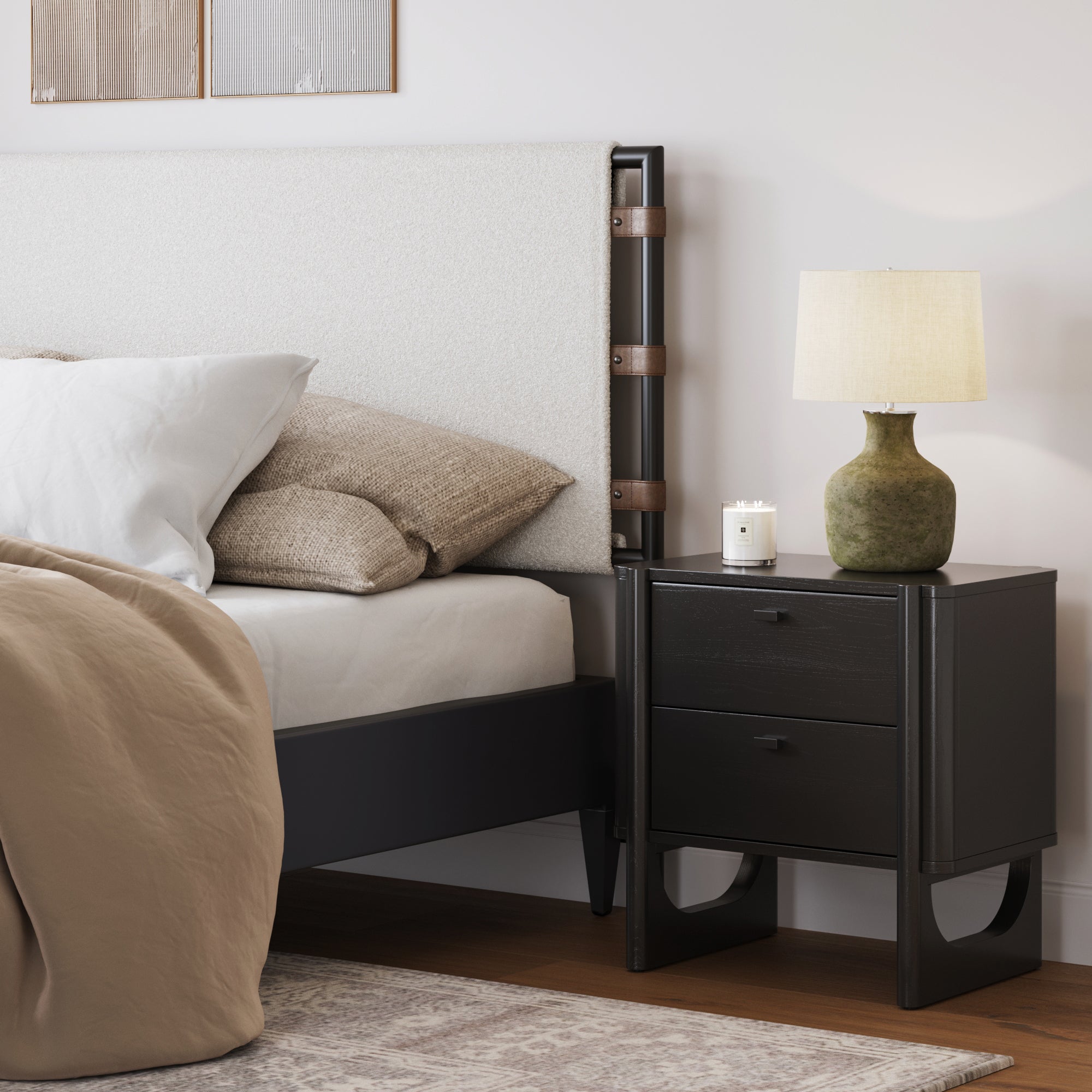 A modern bedroom features a bed with beige bedding and pillows, the Nathan James Mid Century Modern 2-Drawer Solid Wood Nightstand in black, a green ceramic lamp with a beige shade, a candle, and light wall art above the upholstered headboard.