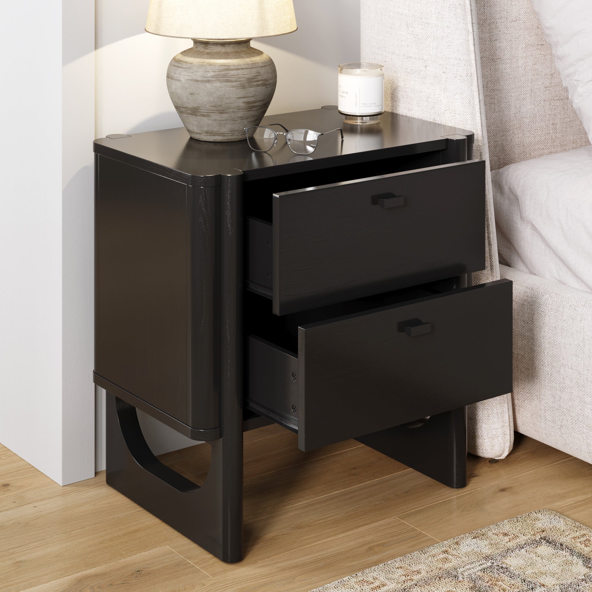 A modern black nightstand with two open drawers stands beside a bed. A small lamp, eyeglasses, and a candle rest on top. The nightstand has a unique cut-out leg design and sits on a wood floor.