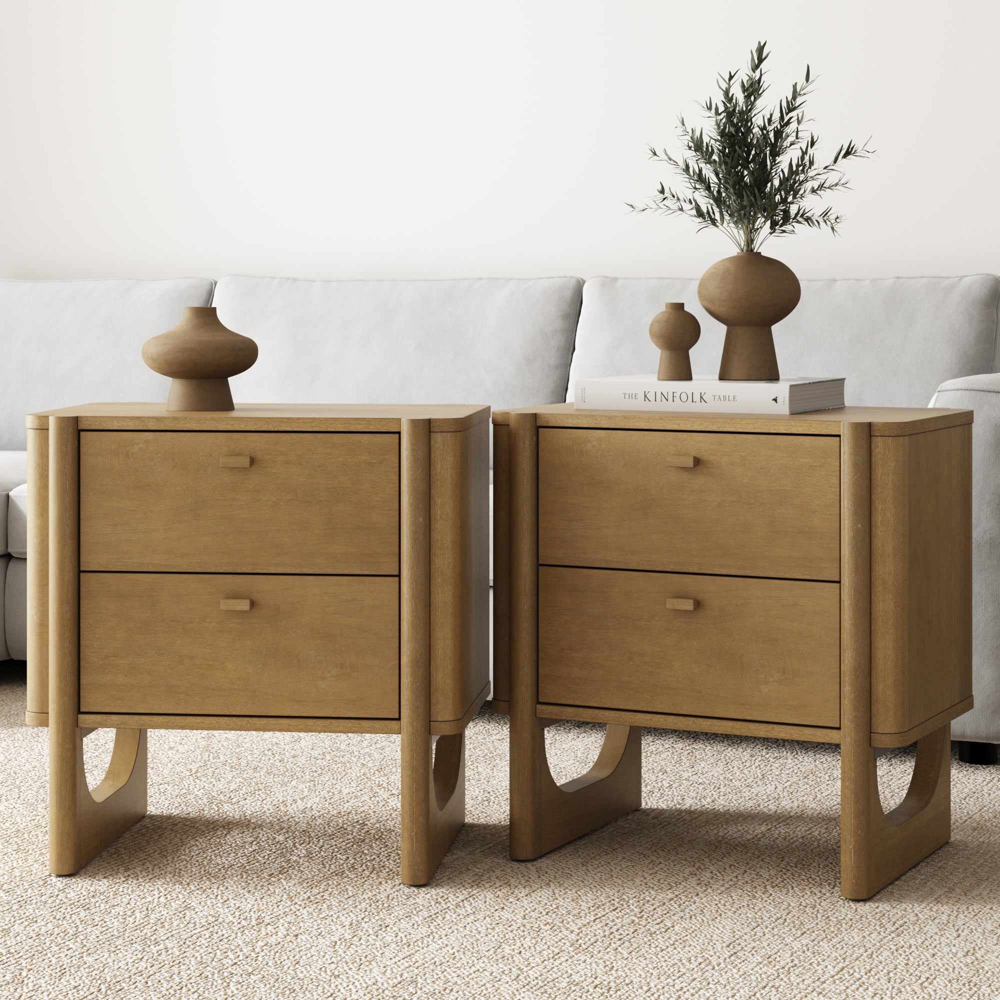 Two Nathan James Mid Century Modern 2-Drawer Solid Wood Nightstands in light brown sit side by side: one holds two ceramic vases, the other a plant, vase, and books. A light sofa and neutral carpet complete the serene setting.