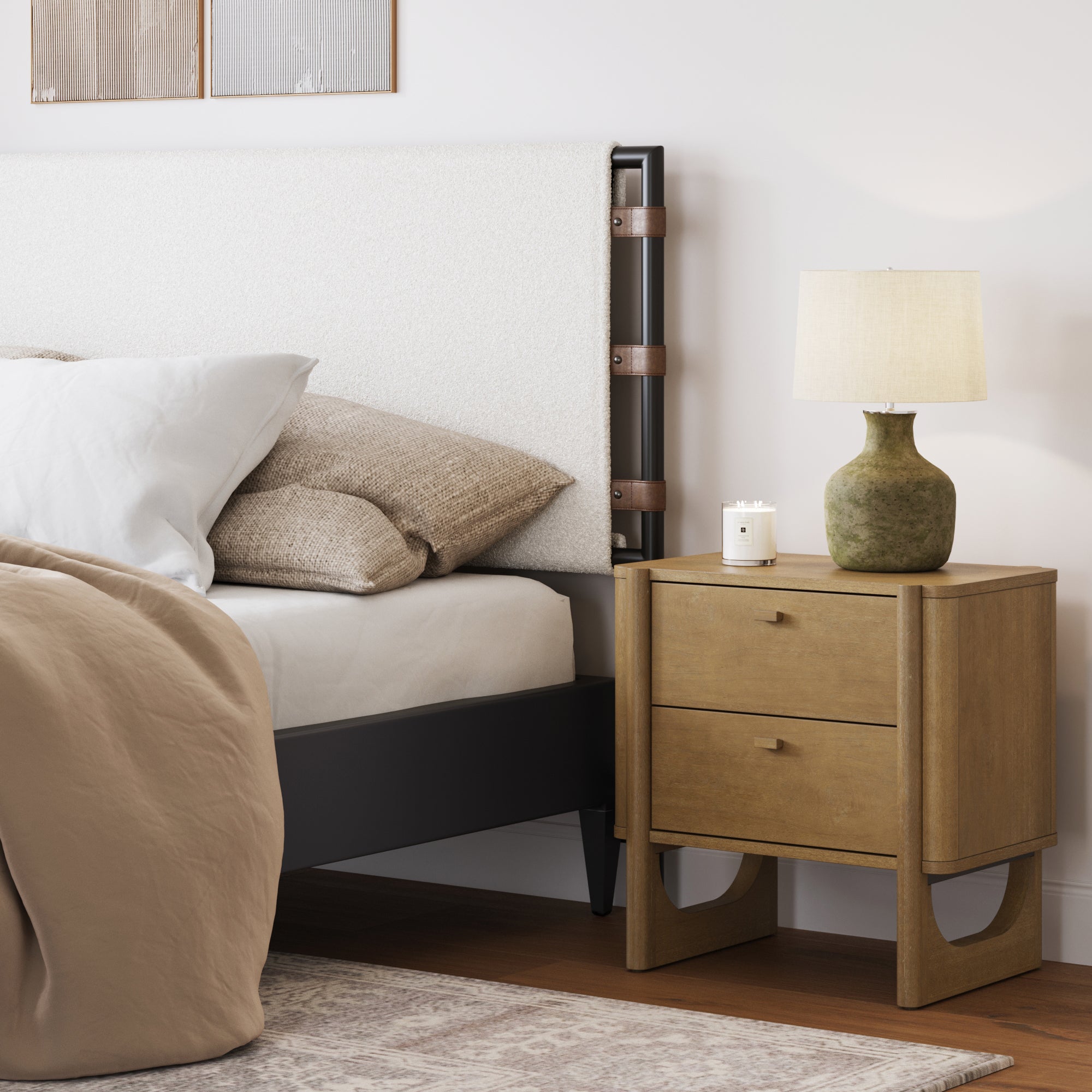 A modern bedroom scene featuring the Nathan James Mid Century Modern 2-Drawer Solid Wood Nightstand in light brown, topped with a green ceramic lamp and candle, beside a bed with beige bedding and a white upholstered headboard.