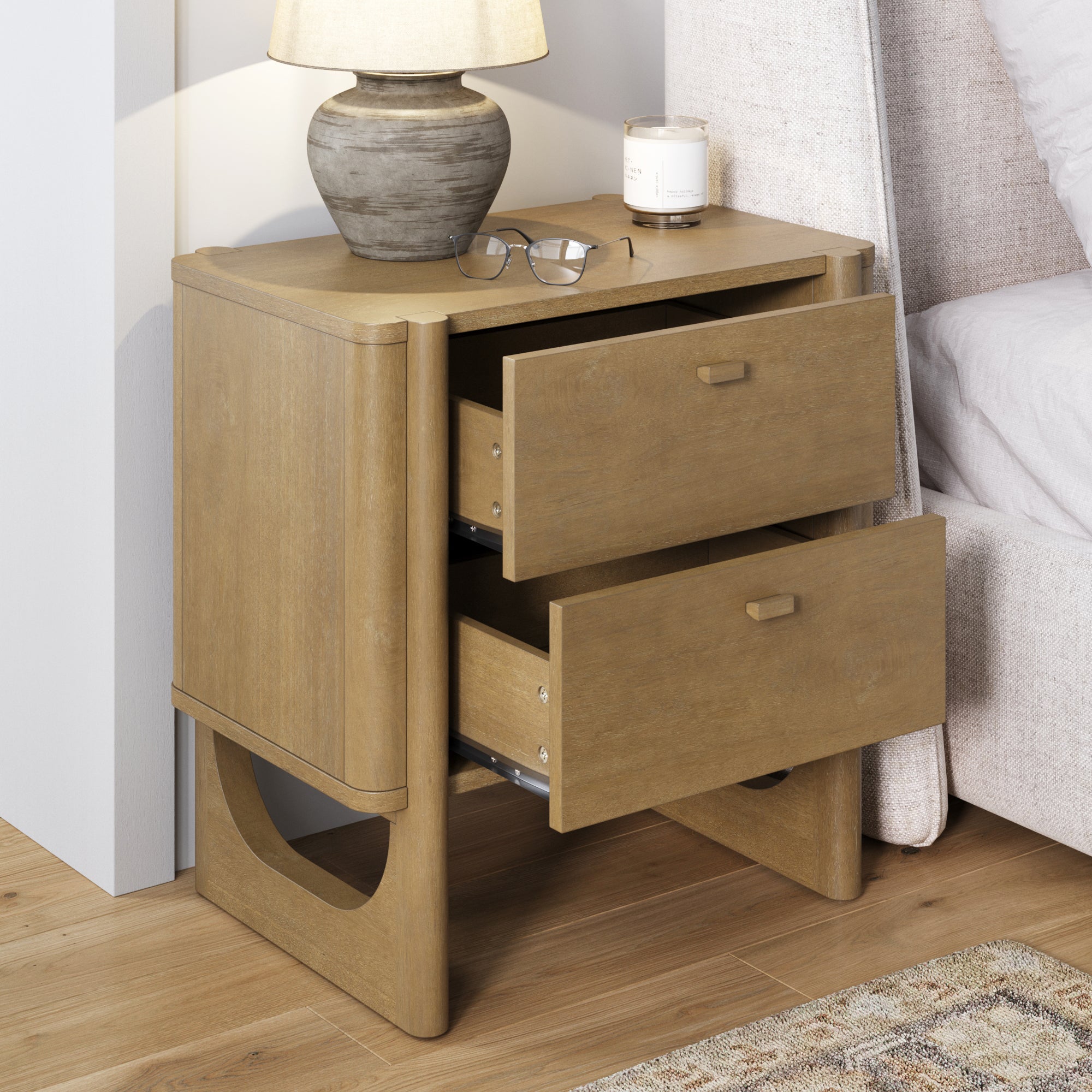 A wooden bedside table with two open drawers, a beige lamp, eyeglasses, and a candle on top, positioned next to a bed with a beige fabric headboard on a wooden floor.