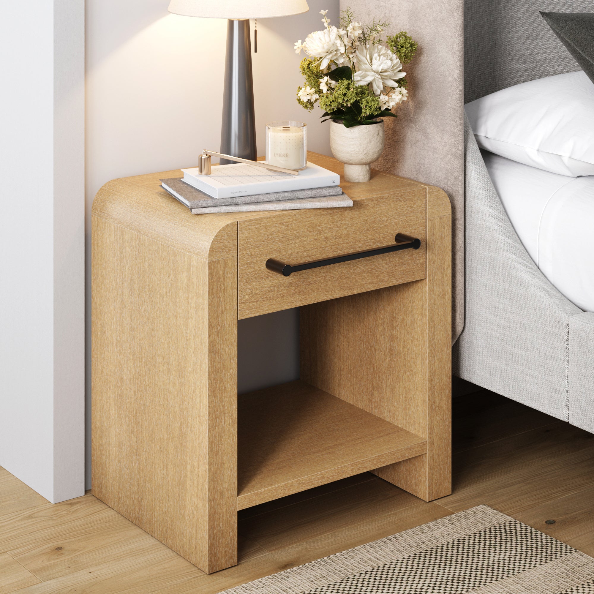 The Nathan James Modern 1-Drawer Nightstand with Metal Handle in Light Brown offers ample storage with a drawer and open shelf—ideal for keeping books, a lamp, or flowers handy next to your bed on a wooden floor.