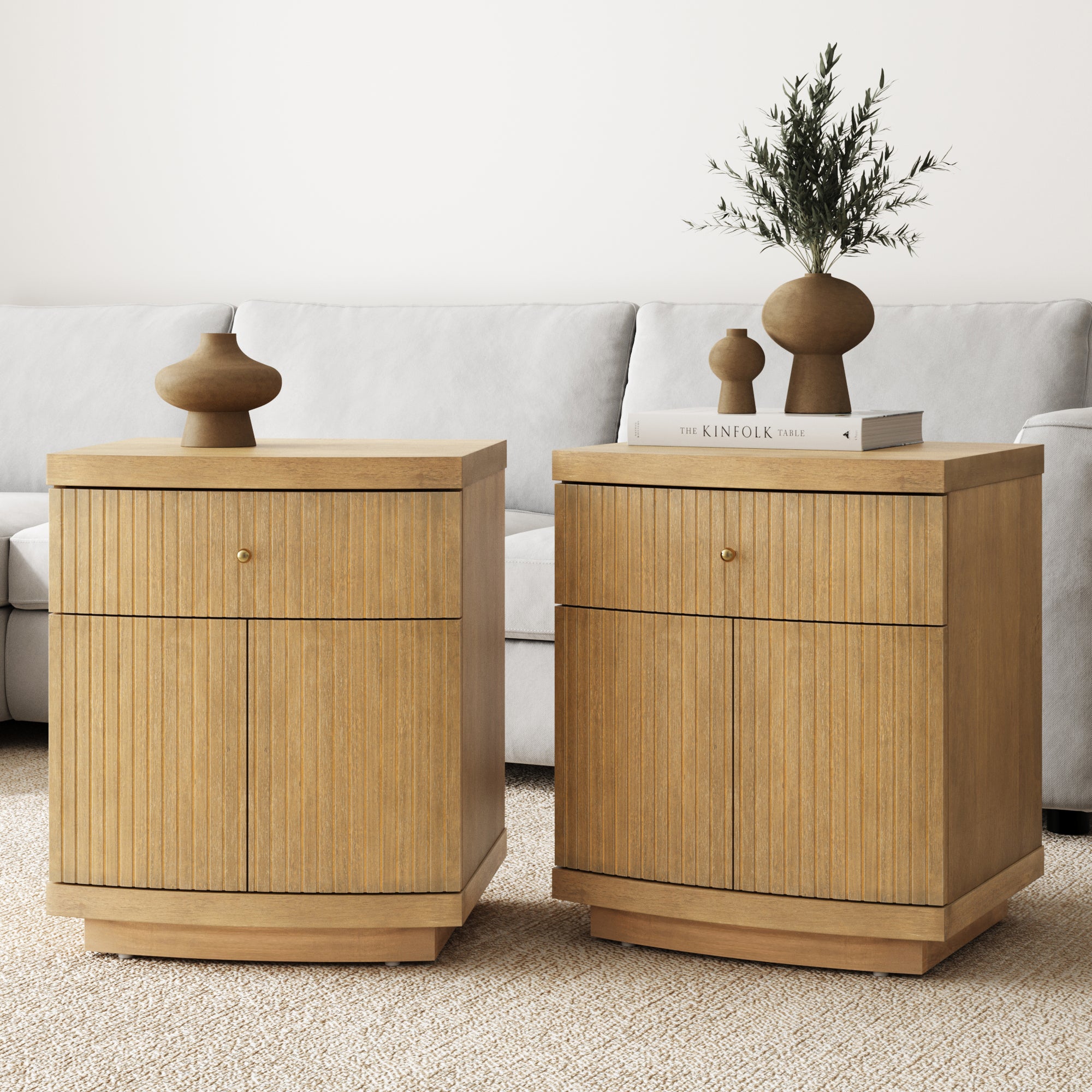 Two Nathan James Fluted Nightstands with drawers and cabinet storage in light brown wood veneer stand on a beige carpet before a light gray sofa. One displays two vases, books, and a green branch in a vase. Set of 2.