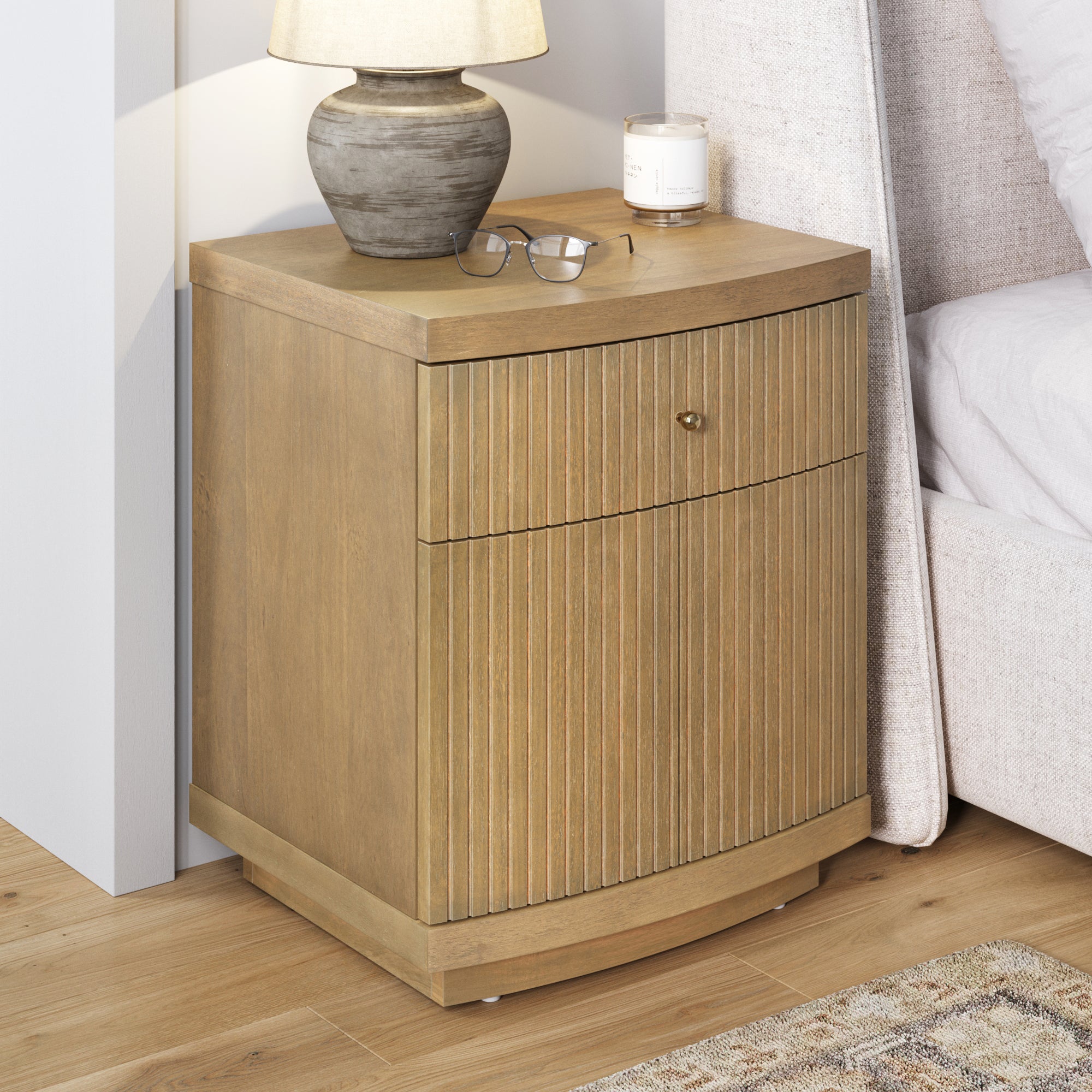 The Nathan James Fluted Nightstand with Drawer and Cabinet Storage in Light Brown features vertical grooves. It sits beside a beige upholstered bed, topped with a gray lamp, eyeglasses, and candle for a modern look.