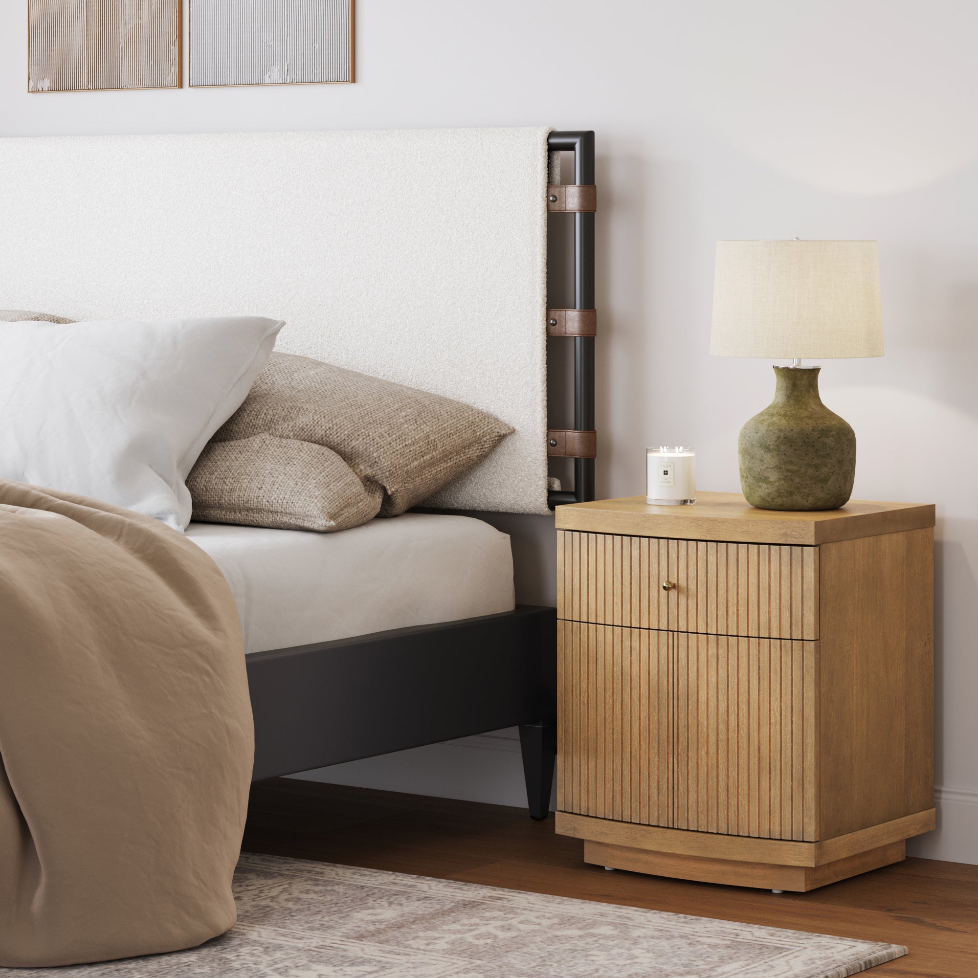 A cozy bedroom corner with a beige bed, white and tan pillows, a Nathan James Fluted Nightstand with Drawer and Cabinet Storage in light brown, a green ceramic lamp with a beige shade, a small candle, and a soft rug on hardwood floors.