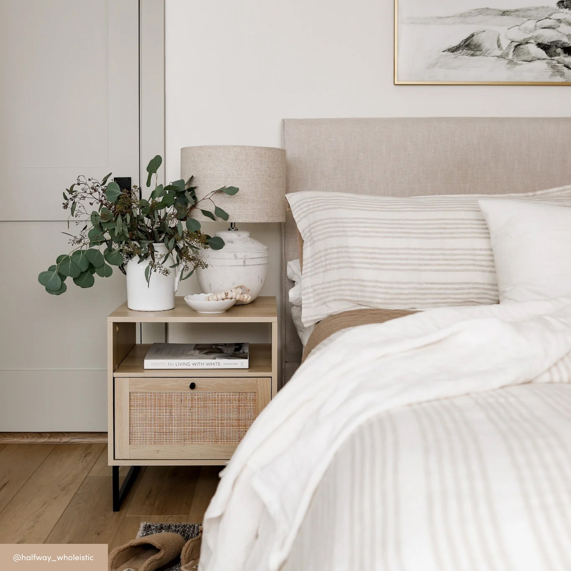 A cozy bedroom with a beige upholstered bed, striped bedding, and a light wood nightstand. The nightstand holds a ceramic lamp, a plant in a vase, and stacked books. The room has soft, neutral tones and a calm, minimalist style.