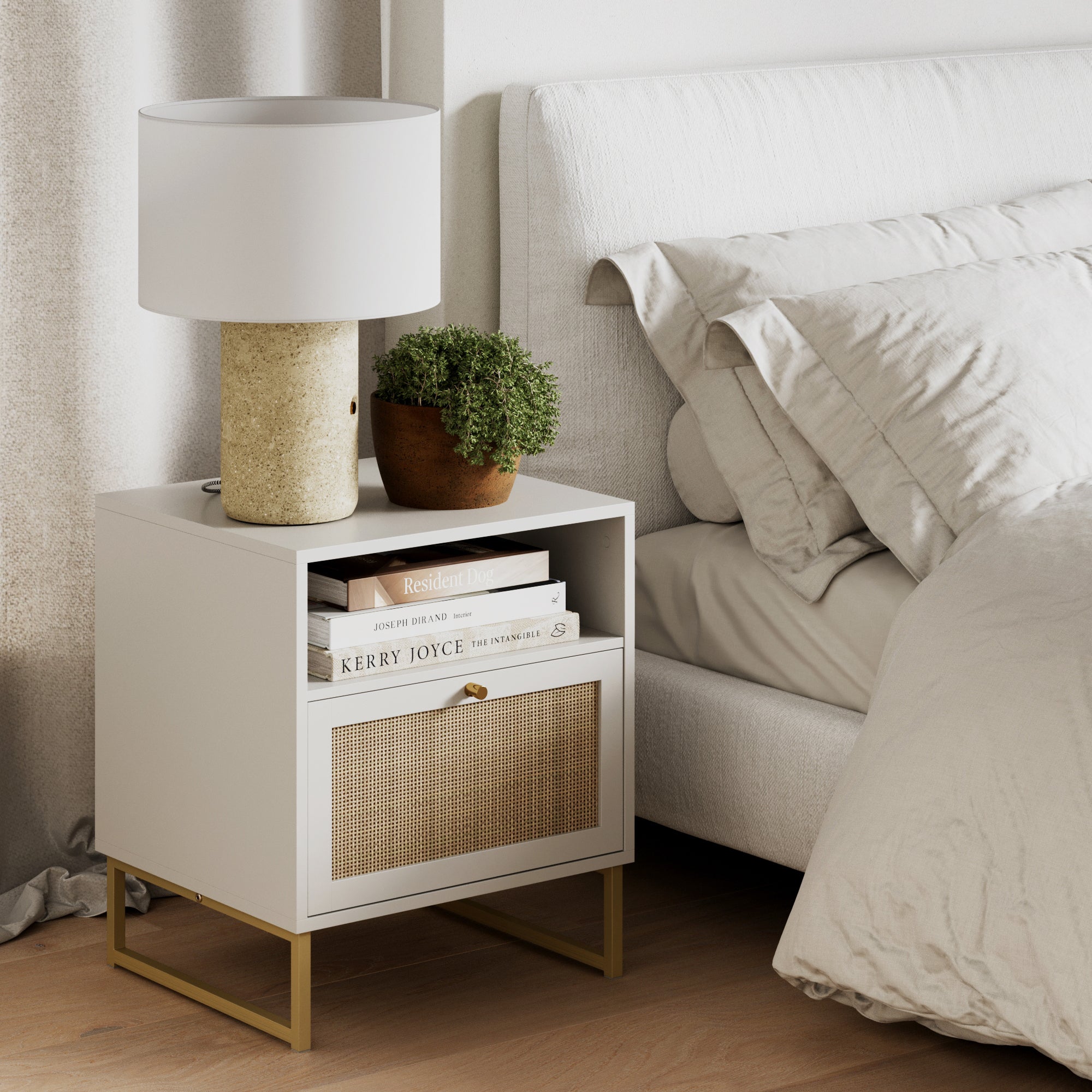 A modern white nightstand with a beige lamp, a small potted plant, and stacked books beside a bed with cream bedding in a minimal, cozy bedroom setting.