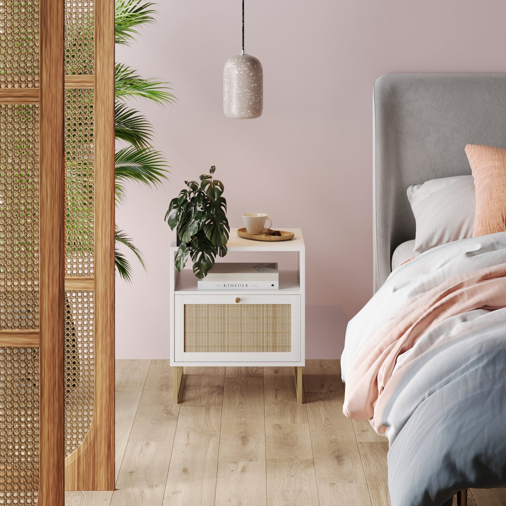 A cozy bedroom features the Nathan James Rattan & Wood Storage Nightstand in white, topped with a plant, book, teacup, and plate. A pendant light hangs overhead, while boho accents like a wooden screen and greenery complement the pink wall.