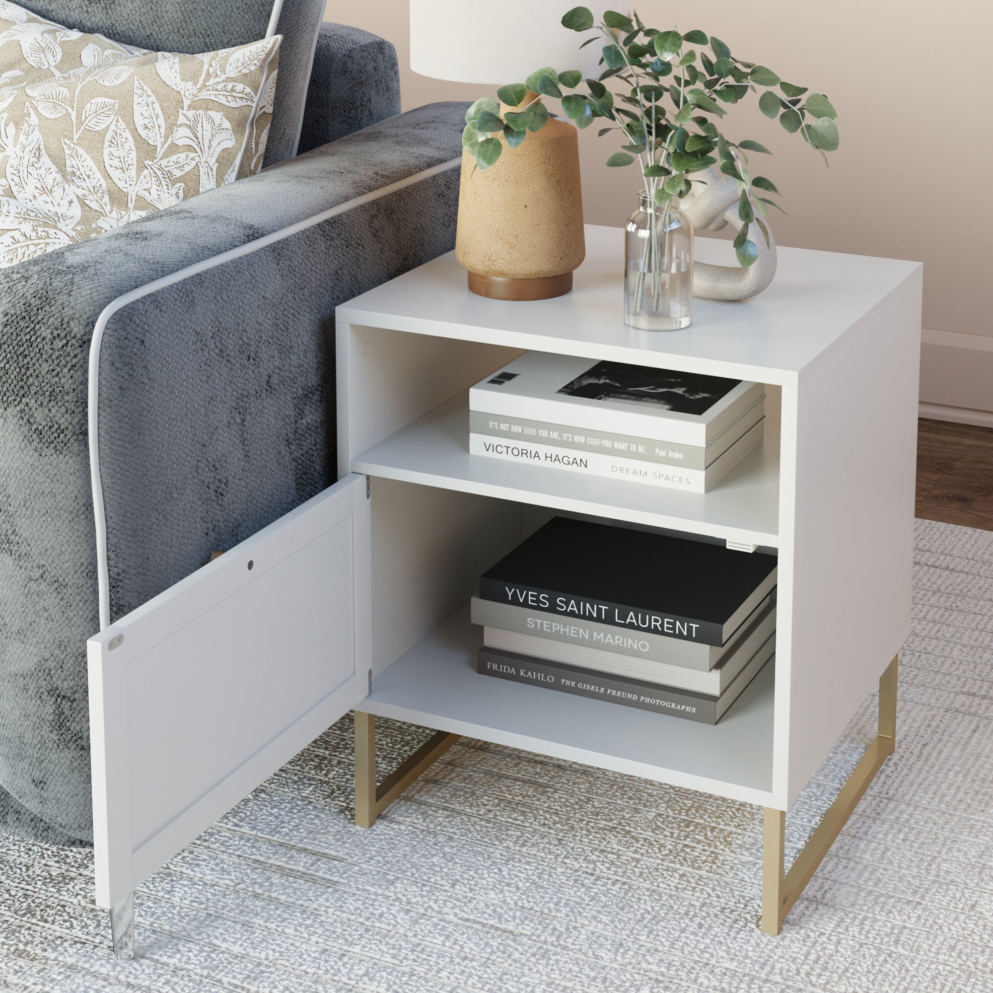 A white side table with gold legs sits beside a gray sofa. On top are a tan vase with a plant and a clear vase with greenery. The shelves hold books, with one cabinet door open. A patterned rug is underneath.