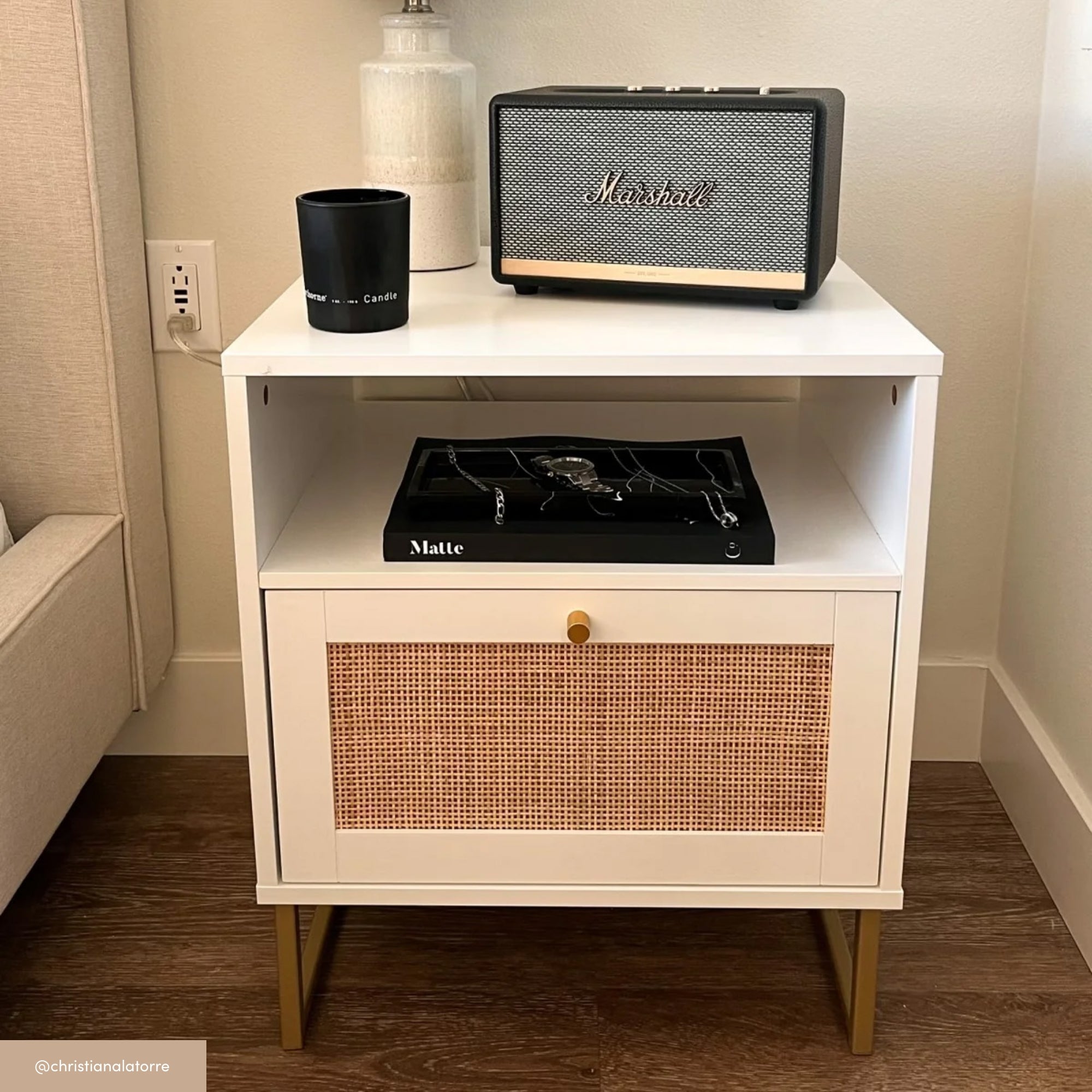 A modern white nightstand with gold legs holds a lamp, a black Marshall speaker, a black candle, and a tray with accessories. The lower compartment features a woven rattan door.