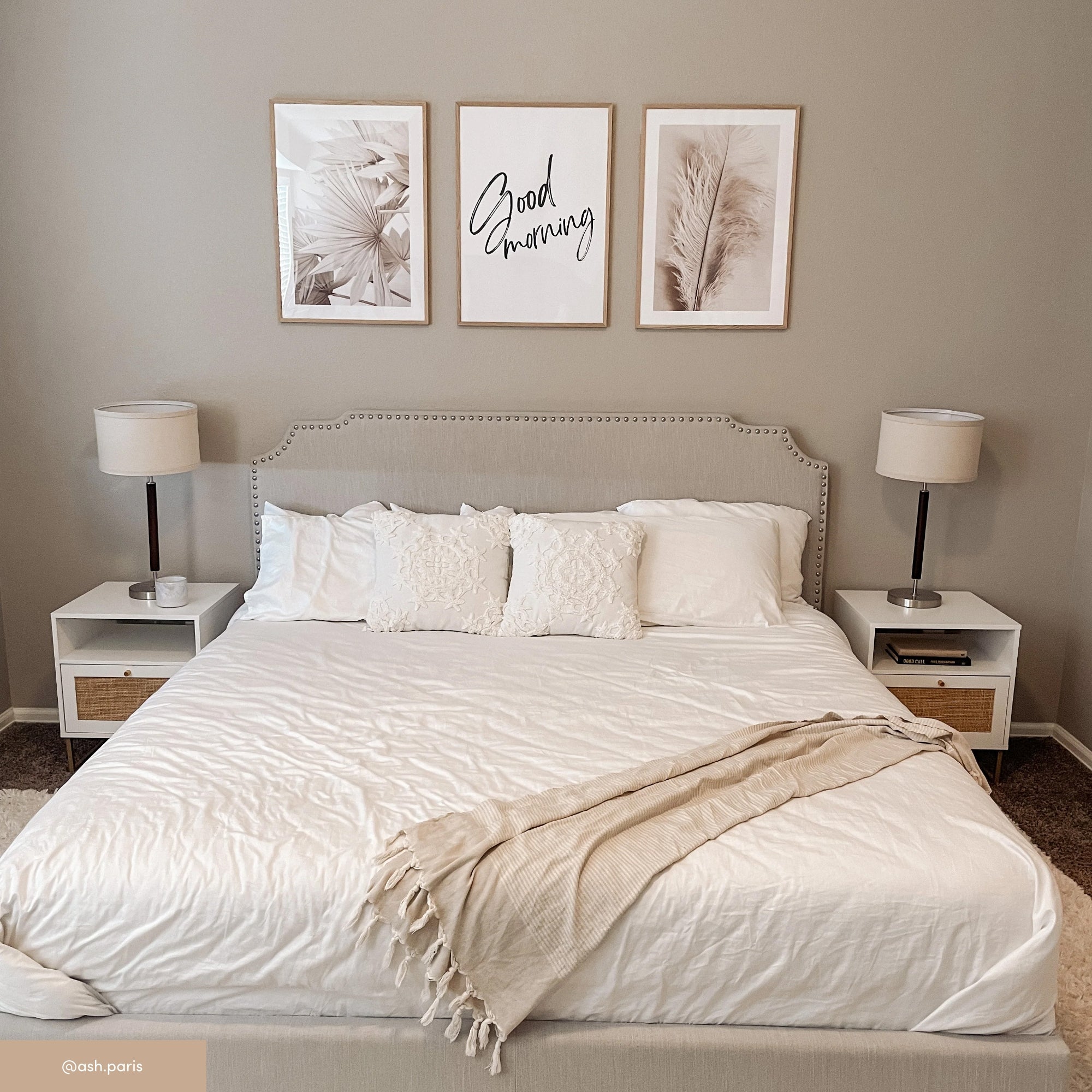 A neatly made bed with white bedding and a beige throw blanket. Two white nightstands with lamps flank the bed. Above the headboard are three framed art prints, one of which says “Good morning.”.