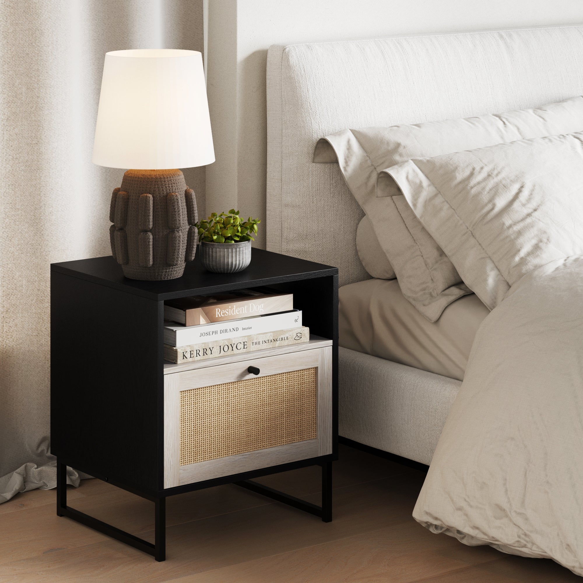 A modern bedside table with a lamp, a small potted plant, and stacked books sits beside a bed with light bedding and pillows on a wooden floor.