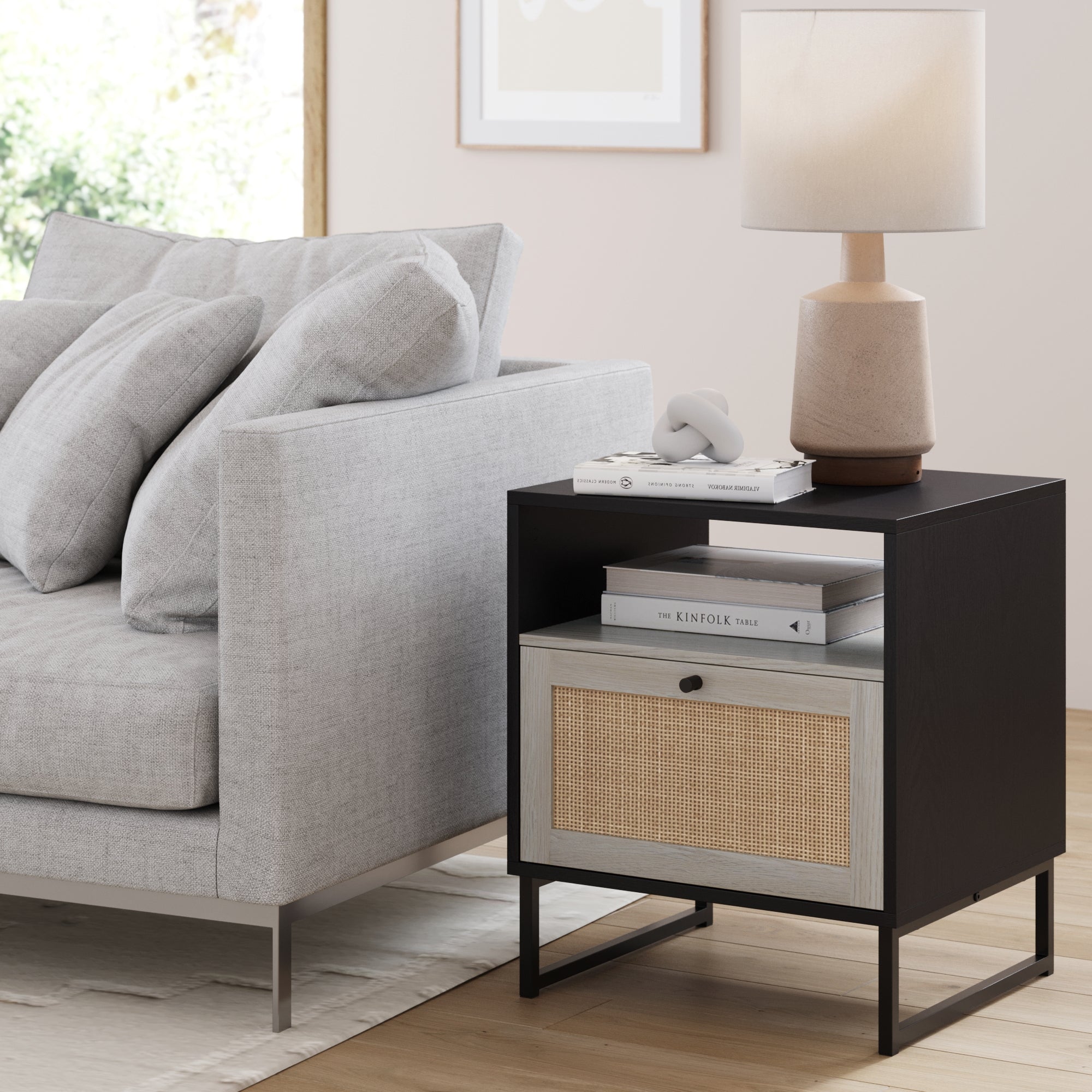 A modern living room features a light gray sofa and the Nathan James Rattan & Wood Storage Nightstand in black, styled with stacked books, decor, and a beige lamp. Sunlight pours in, adding cozy Bohemian flair to the space.