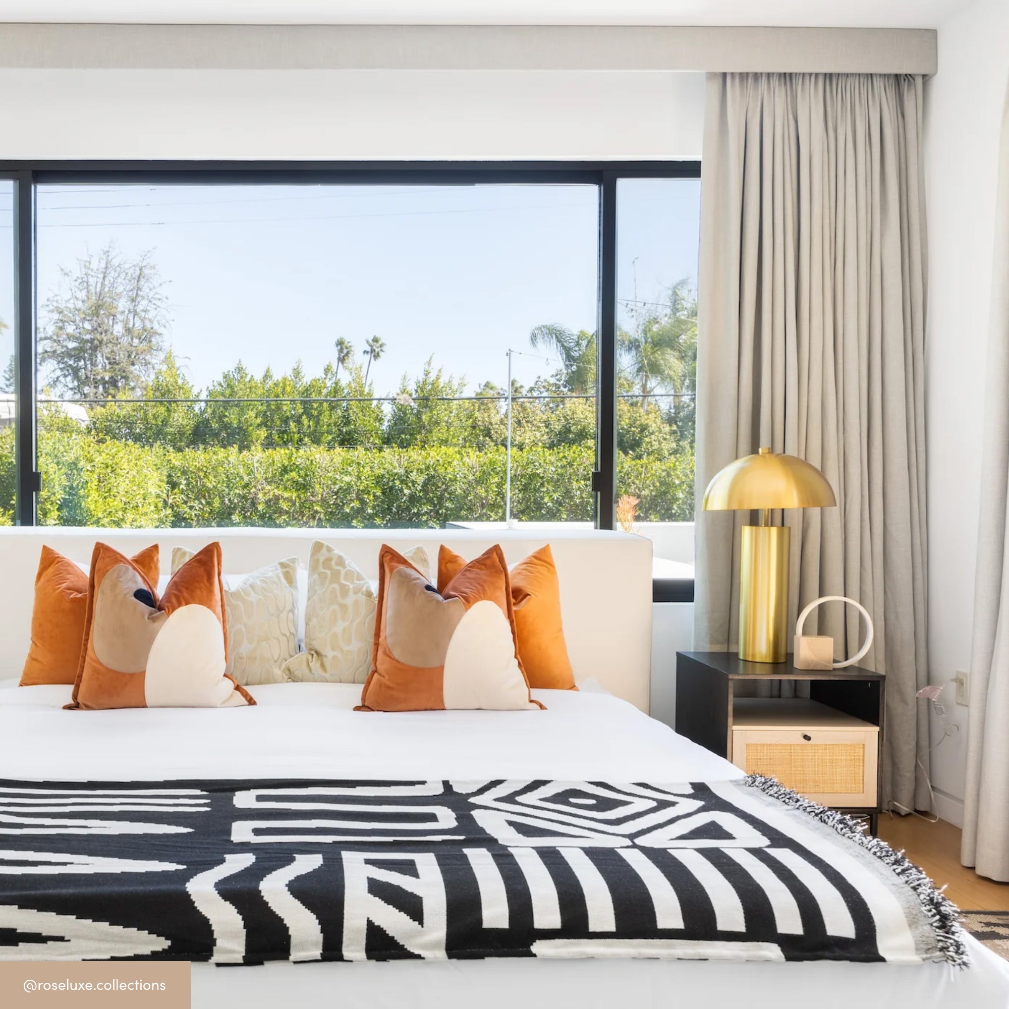 A modern bedroom with a large window, a bed decorated with geometric black and white blankets, orange and cream pillows, light curtains, and a gold lamp on a bedside table. Lush greenery is visible outside the window.