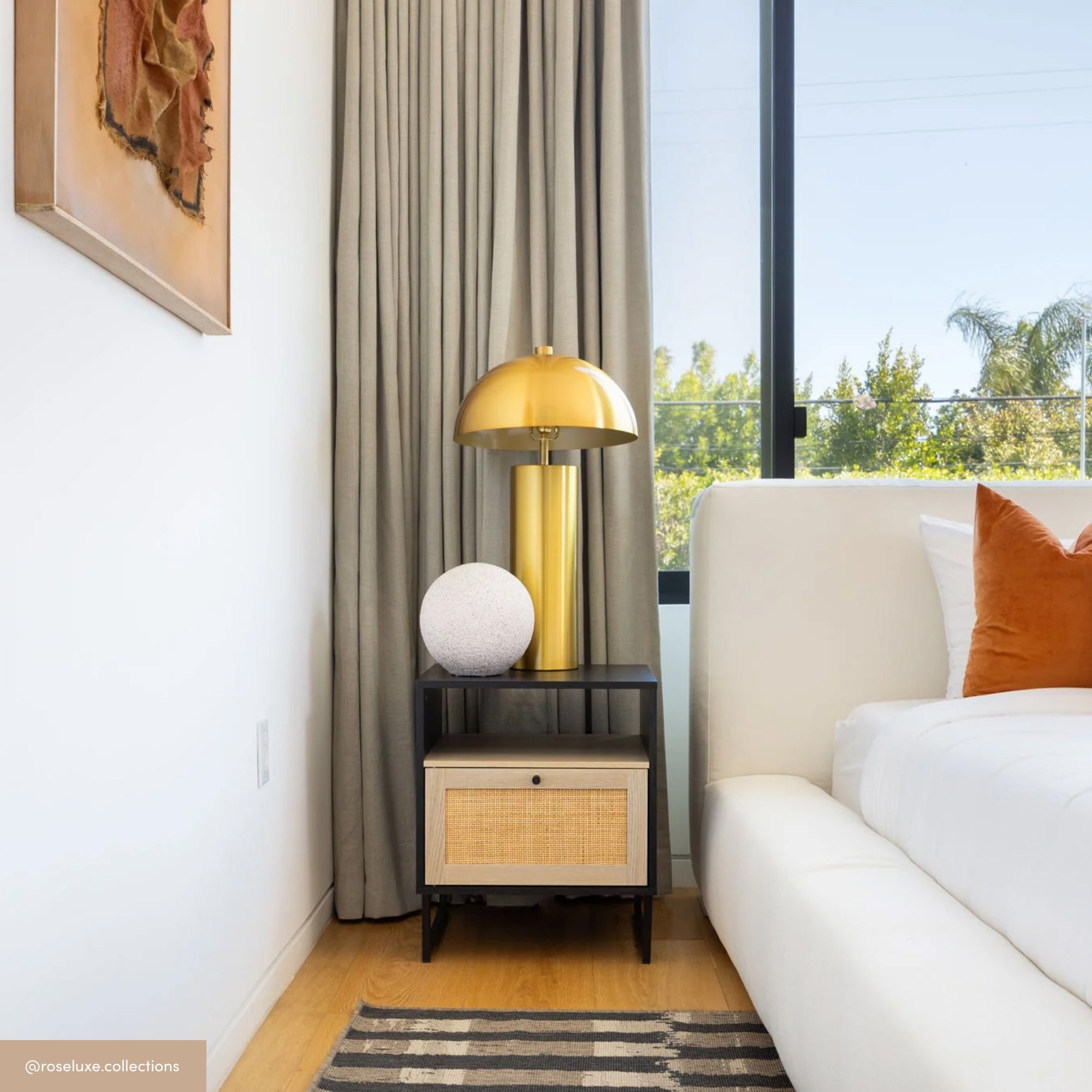 A modern bedroom features a white bed with an orange pillow, a black nightstand holding a round white lamp and a gold lamp, beige curtains, a large window, and a striped rug on a wooden floor.