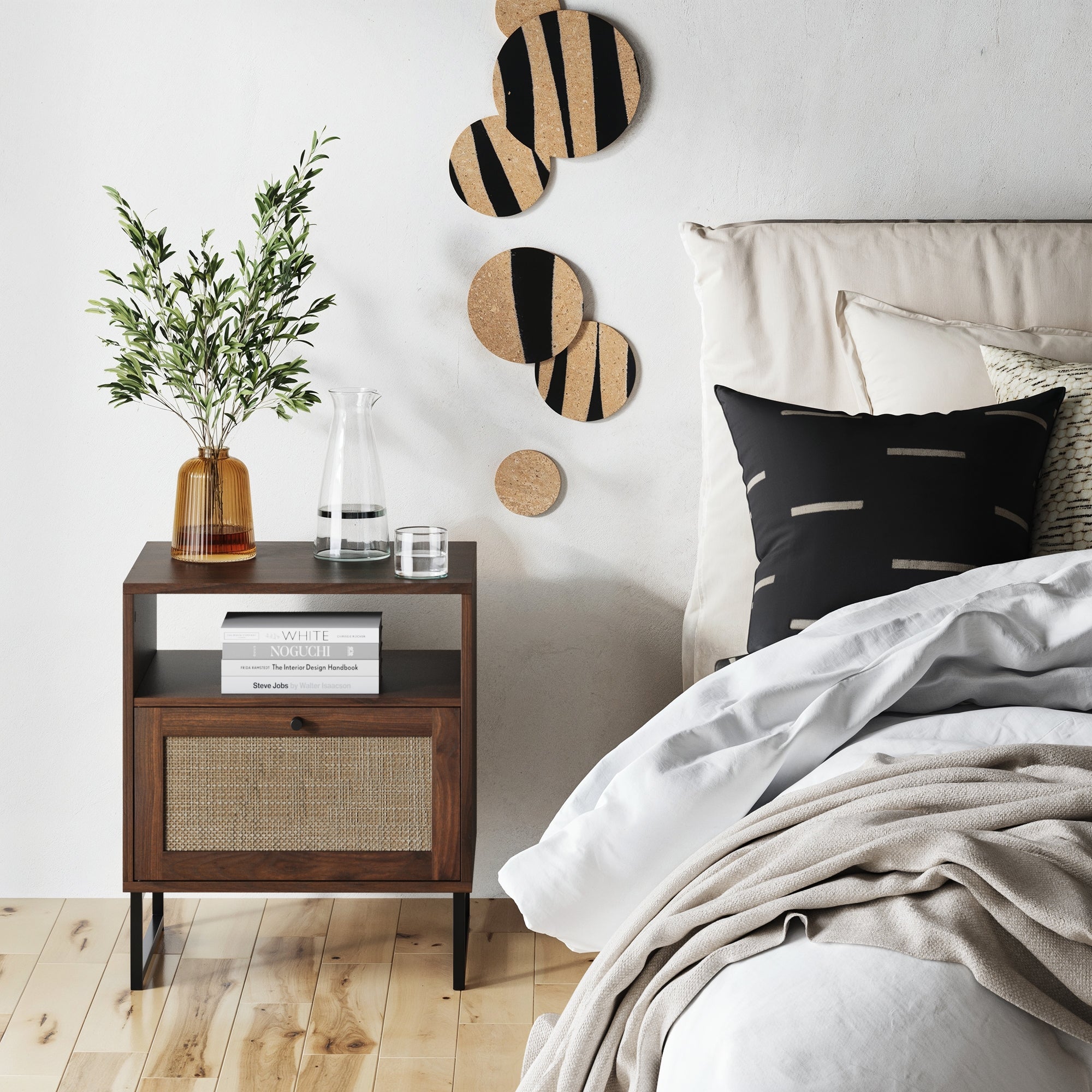 A cozy bedroom scene with a wooden nightstand holding a plant in an amber vase, a glass carafe with a cup, and books; modern abstract wall art and a neatly made bed with black and patterned pillows are nearby.
