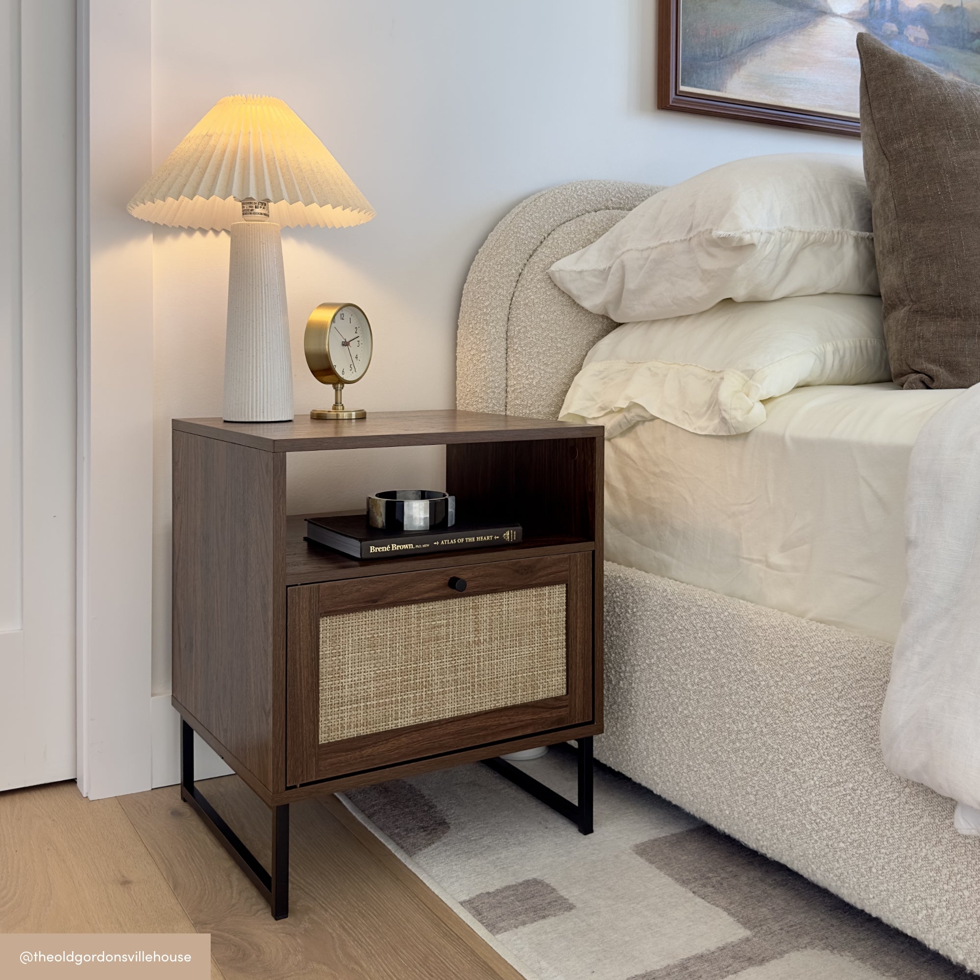 A modern wooden nightstand with a rattan drawer stands beside a bed with cream bedding. On the nightstand are a pleated lamp, a clock, and a small round box. A framed artwork hangs above.