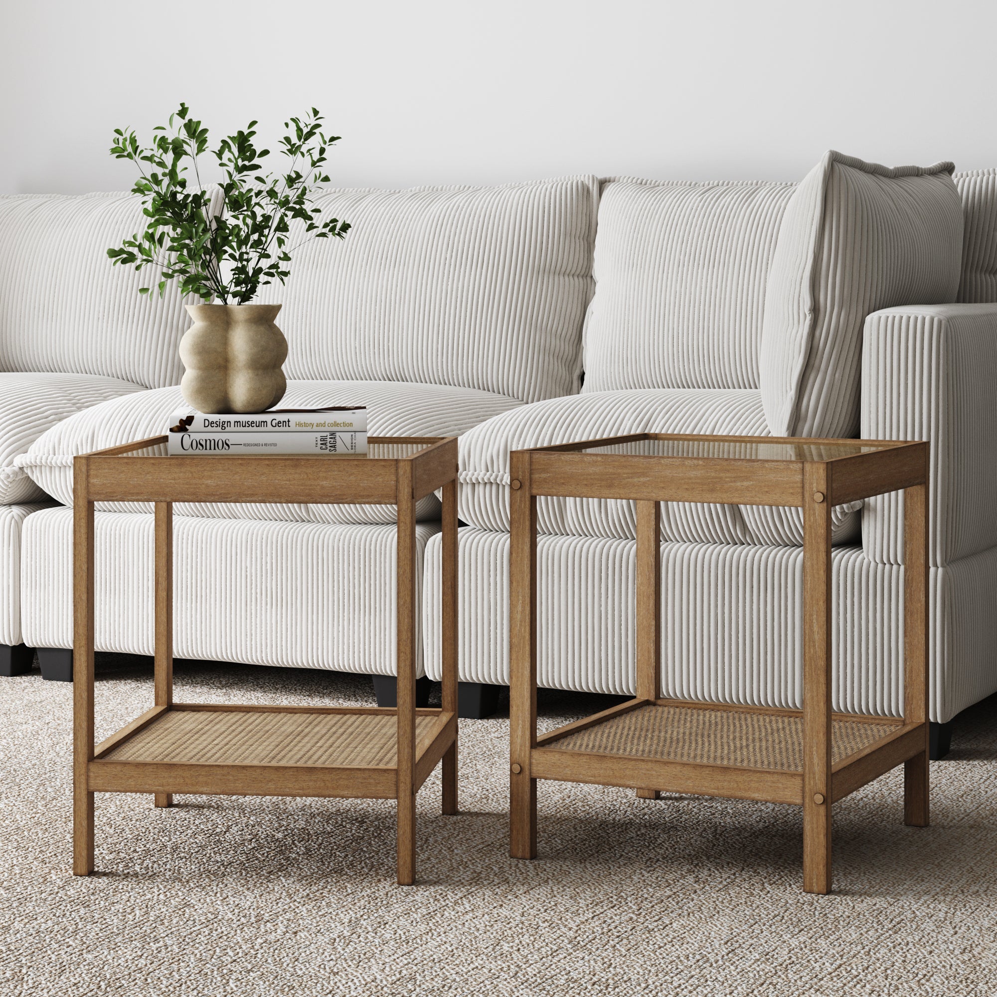 The Nathan James Solid Wood Rattan & Glass End Table Set of 2 features woven shelves and sits in front of a light textured sofa. One table displays a vase with greenery and a book on neutral carpeting—ideal for small, cozy living rooms.