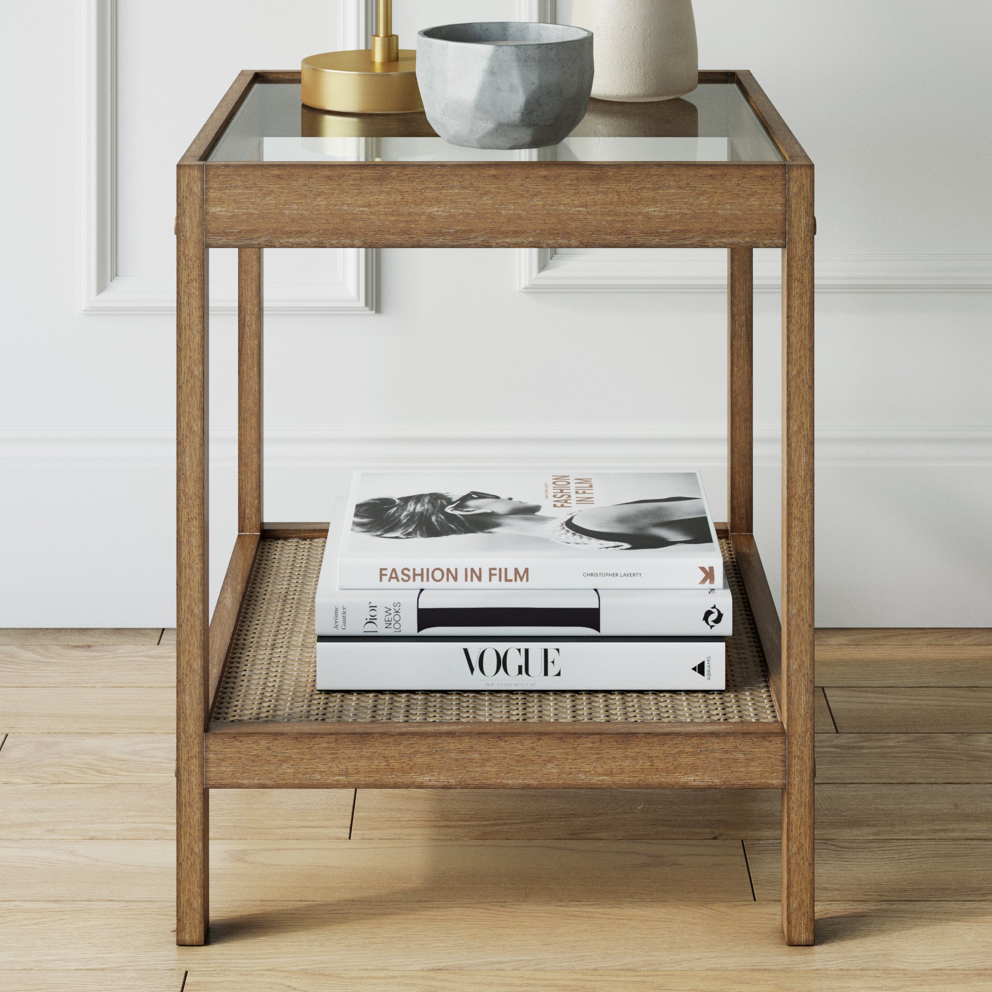 A wooden side table with a glass top holds a decorative bowl and gold object. On the lower shelf, there are two stacked books: Vogue and Fashion in Film with a black-and-white cover image.