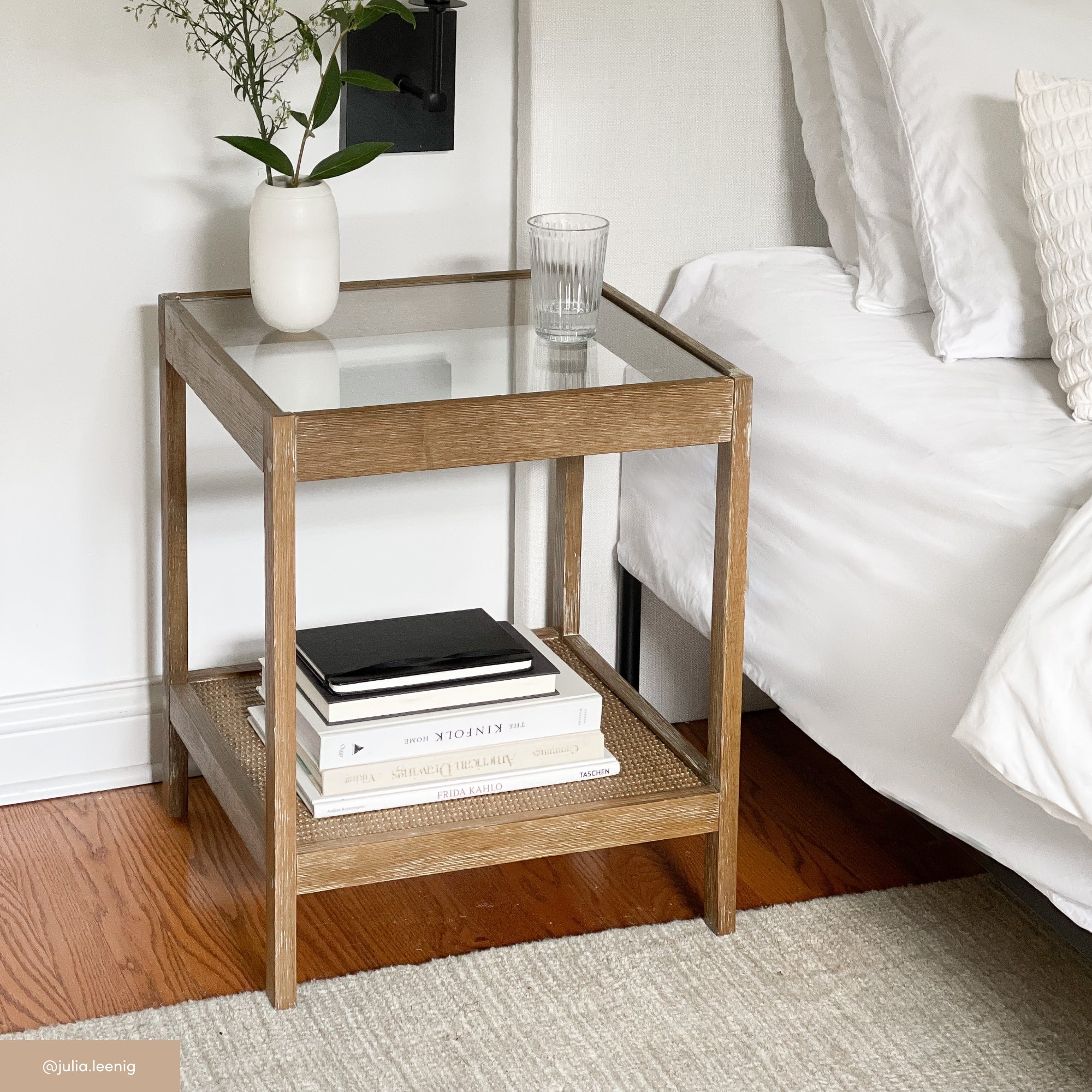A wooden nightstand with a glass top holds a vase with flowers and a glass of water. Books are stacked on the lower shelf. The stand is next to a white bed with pillows, on a hardwood floor.