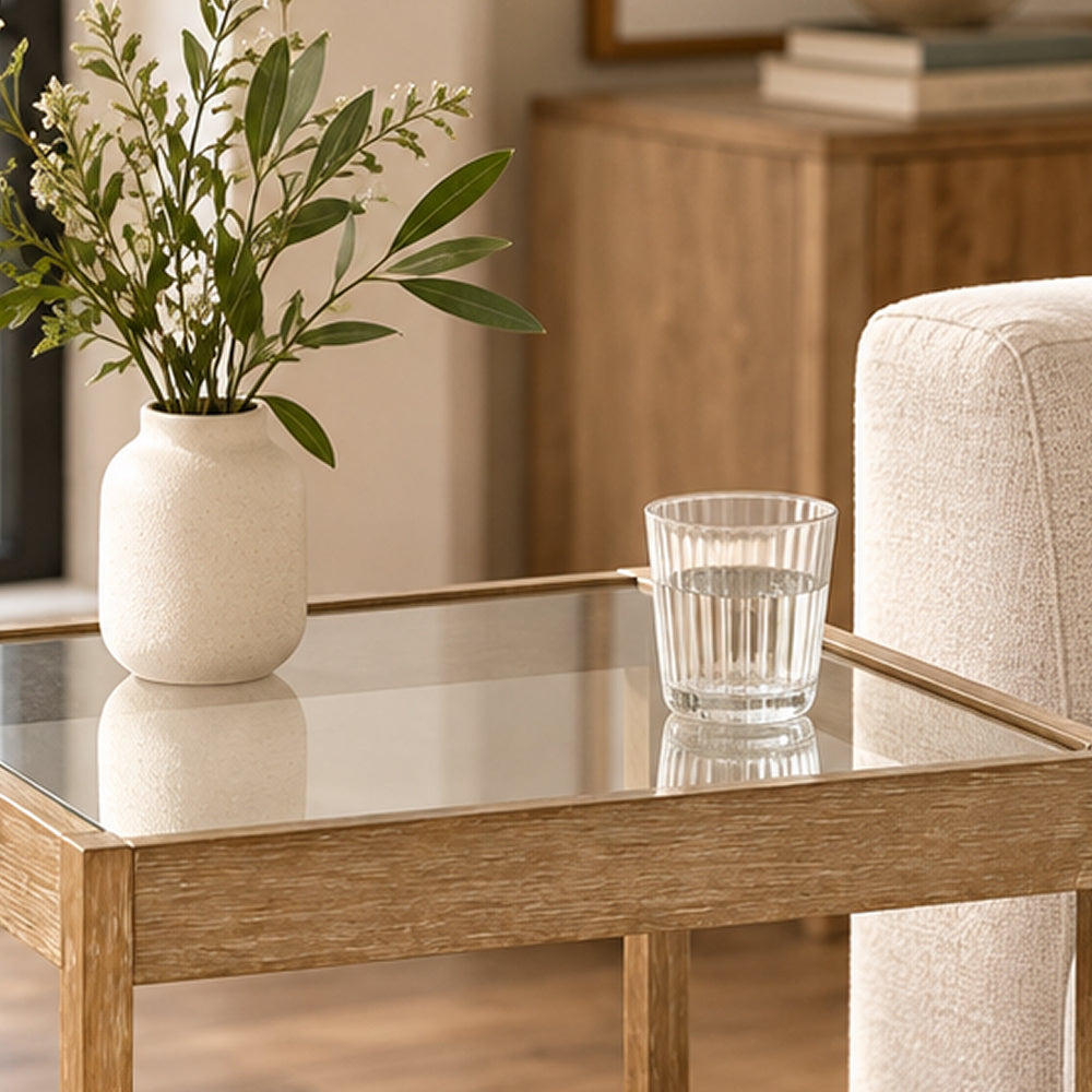 A glass vase with leafy branches and a glass of water on a small wooden side table with a glass top.