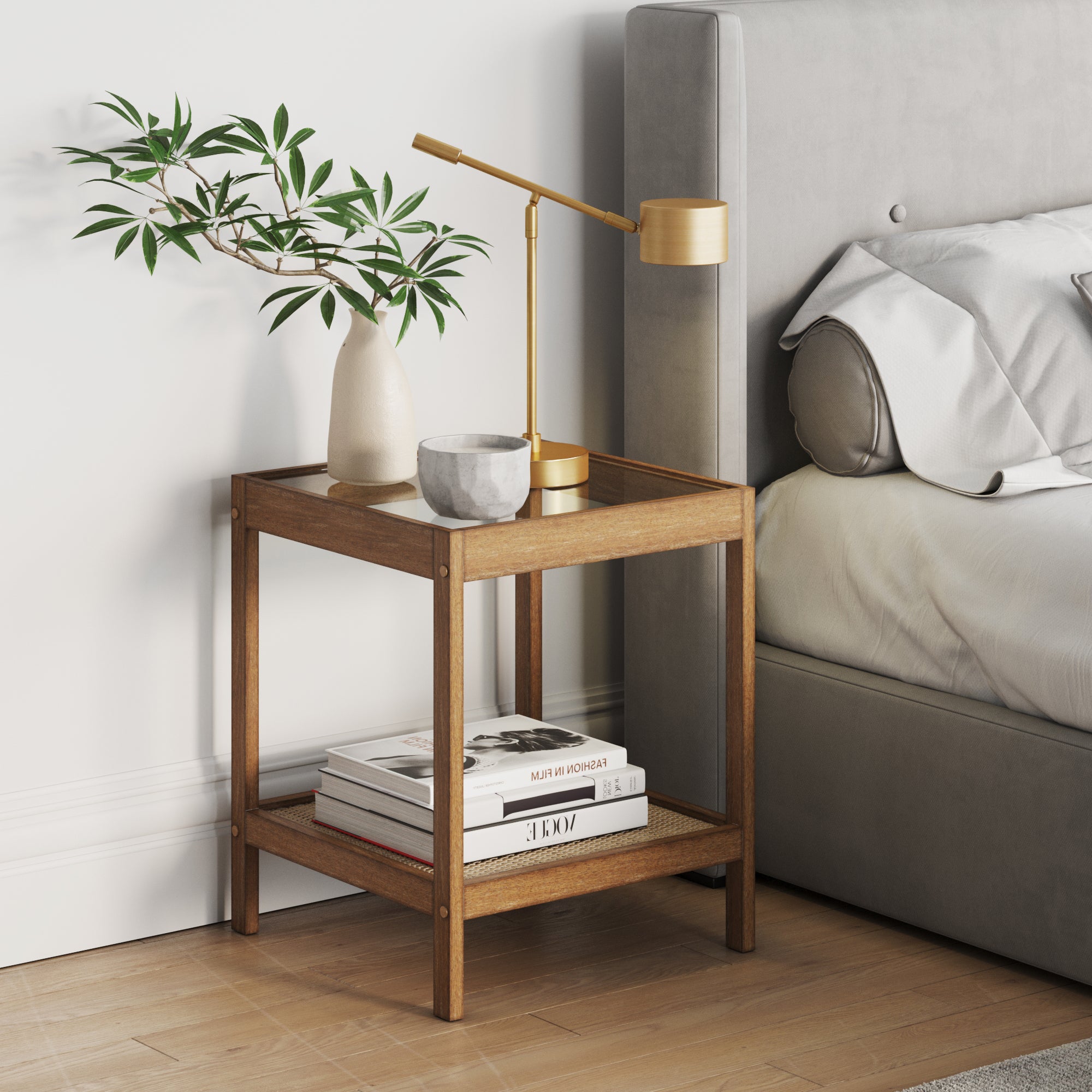 A Nathan James Solid Wood, Rattan & Glass Top End Table holds a potted plant, gray cup, and gold lamp beside a gray upholstered bed. Books sit on its lower shelf in a modern, minimal bedroom.