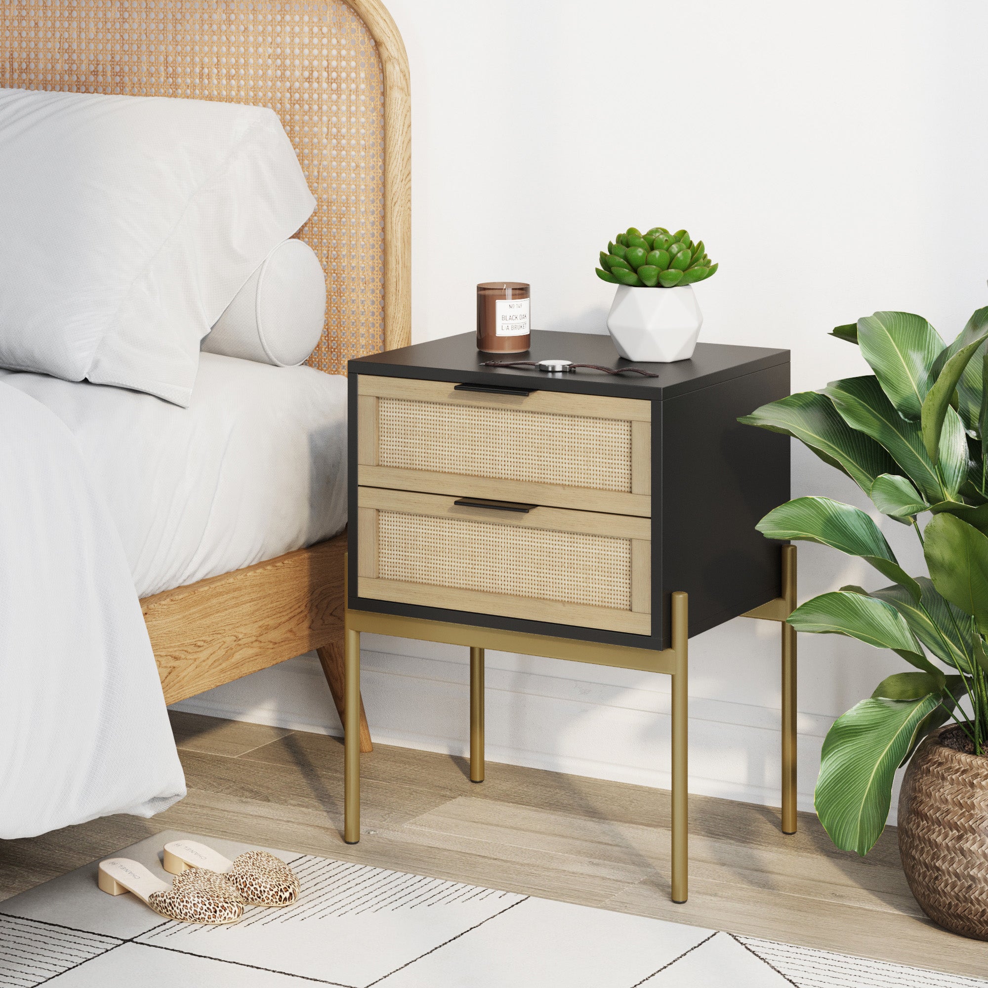 A modern bedside table with rattan drawers, gold legs, and a black top holds a candle, a small dish, and a potted succulent. It stands next to a bed with a wooden frame and a green leafy plant on the floor.