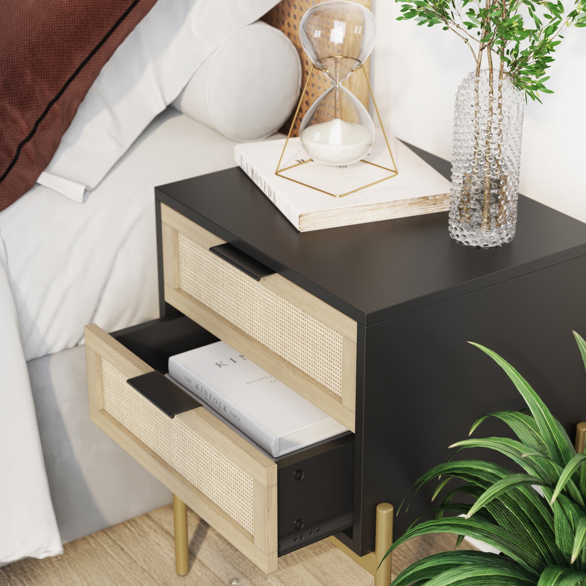 A modern black nightstand with two partially open wooden drawers holds books inside. On top are an hourglass, a textured glass vase with dried flowers, and stacked books. A plant and part of a bed are nearby.