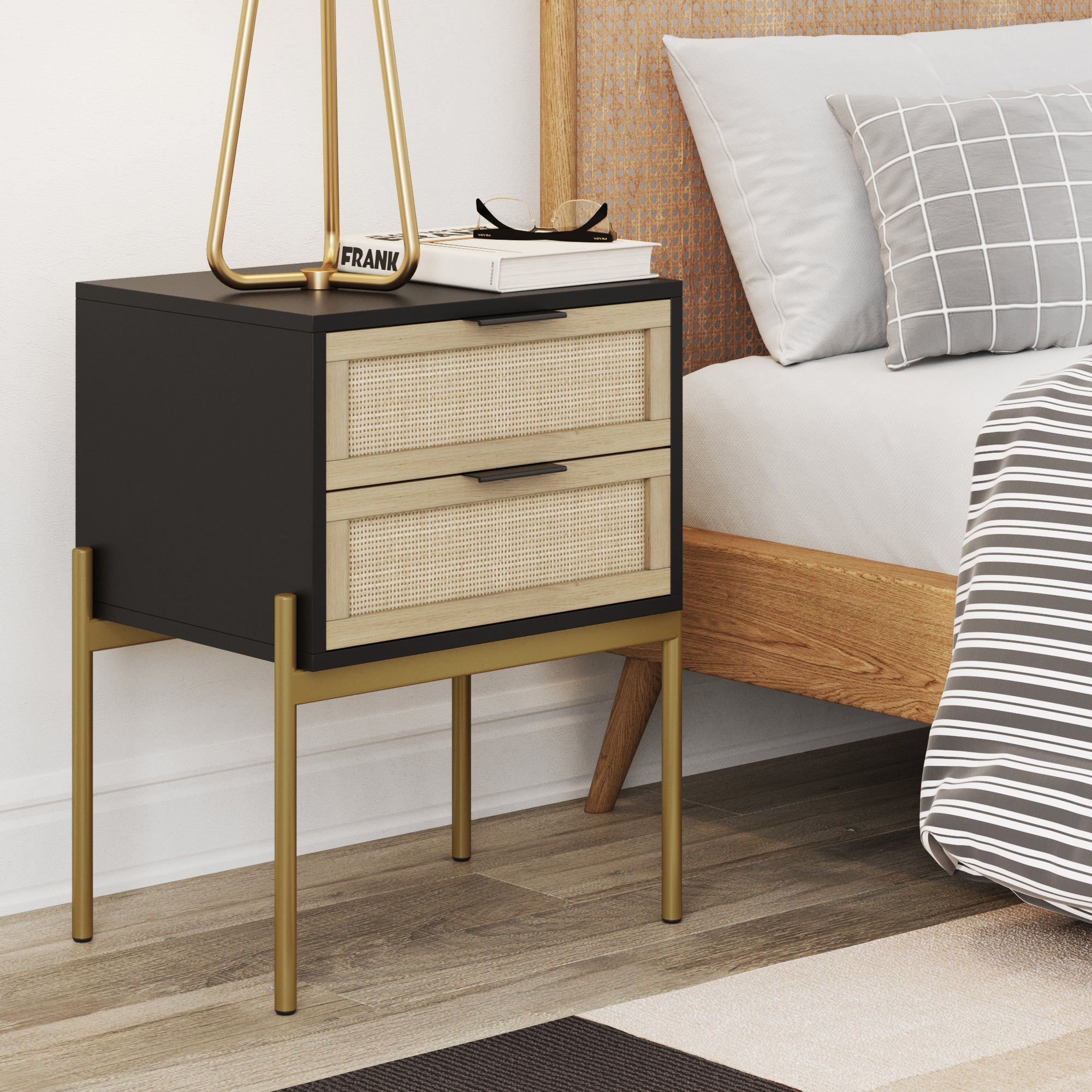 A modern nightstand with black sides, wicker-front drawers, and gold legs stands beside a bed with neutral bedding and a wooden headboard. Items on the nightstand include a lamp and a pair of glasses.