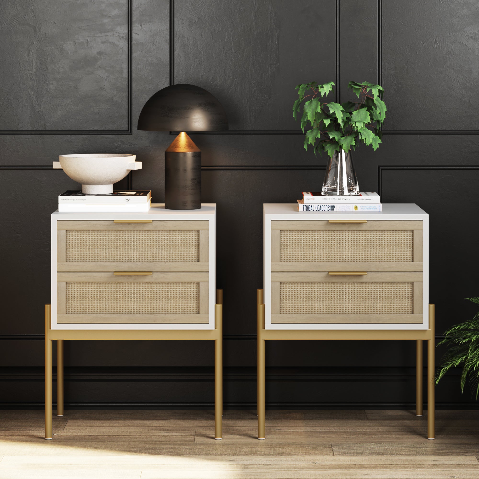 Two modern nightstands with light wood drawers and gold legs sit against a dark paneled wall. One holds a decorative bowl and lamp; the other holds stacked books and a leafy plant in a glass vase.